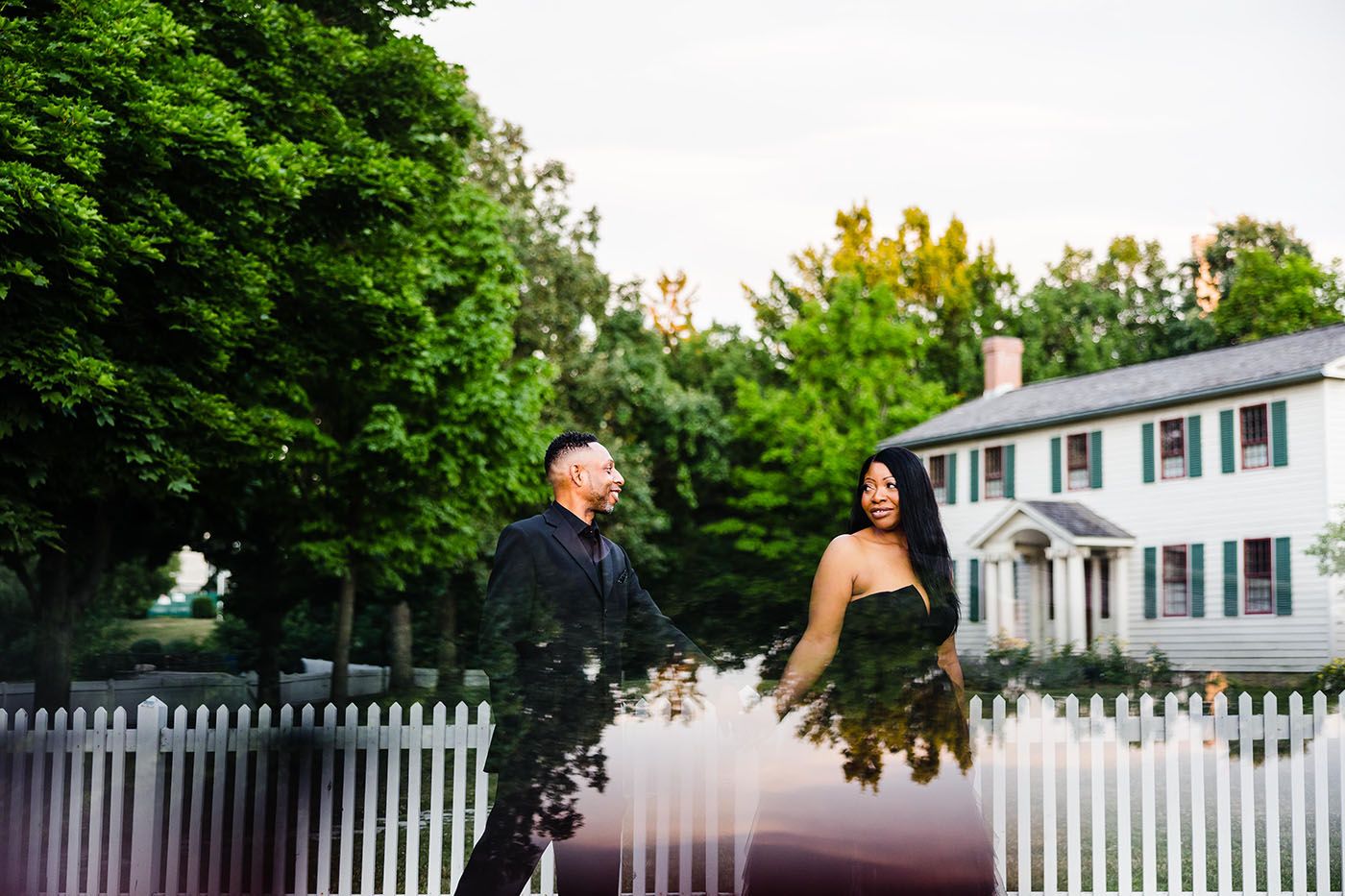 Couple standing together in a garden setting with white picket fence and colonial house in background.