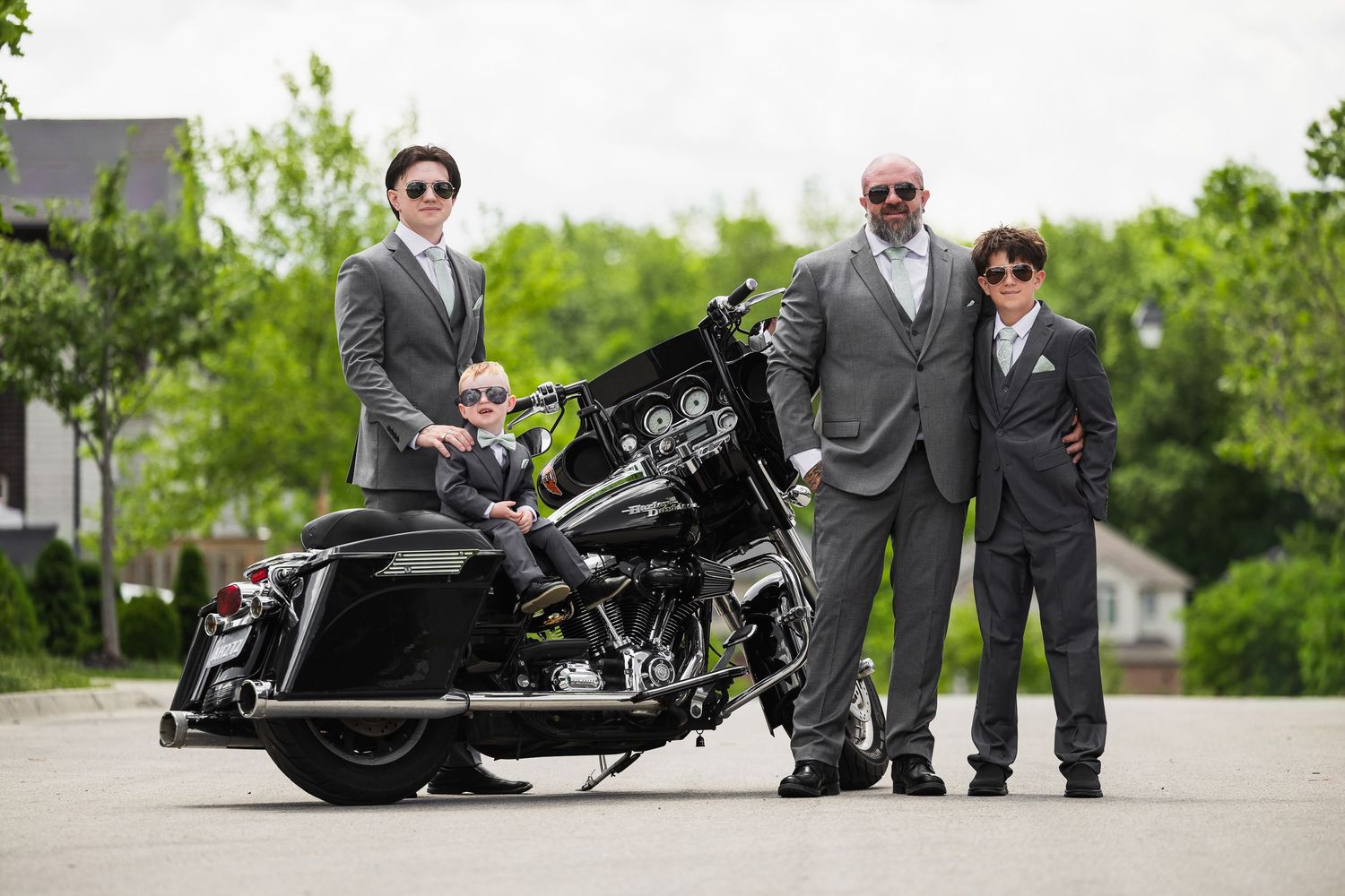 Three males in gray suits posing with a black Harley-Davidson motorcycle outdoors on a sunny day.
