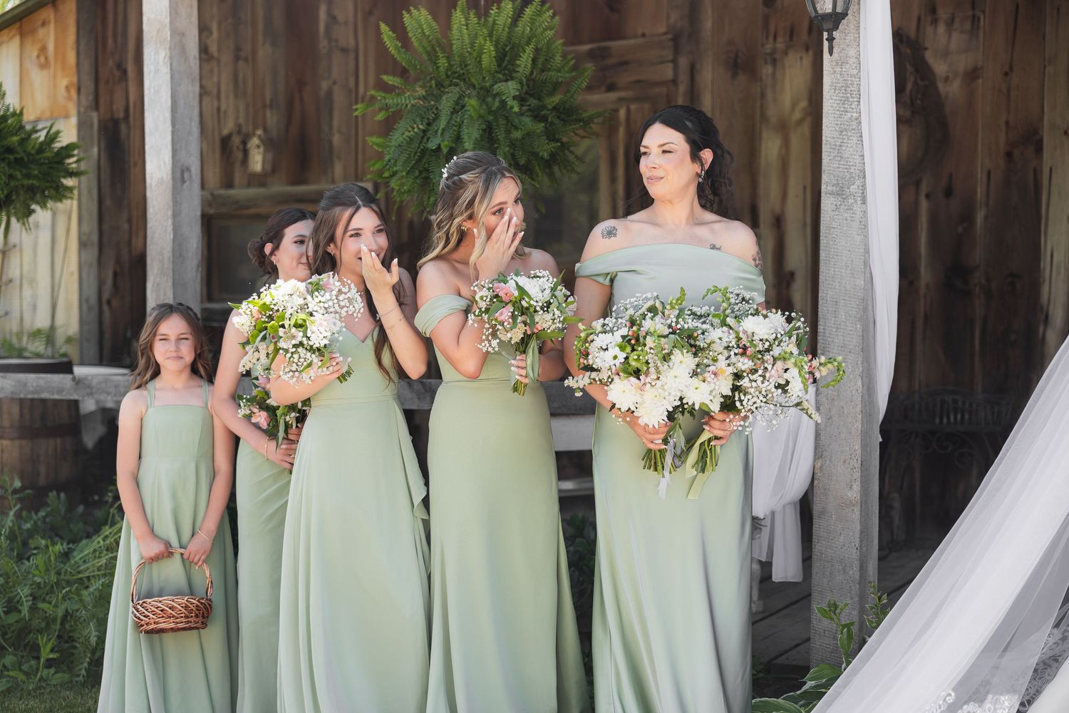 Bridesmaids in sage green gowns holding white bouquets react emotionally outside a rustic barn wedding venue.