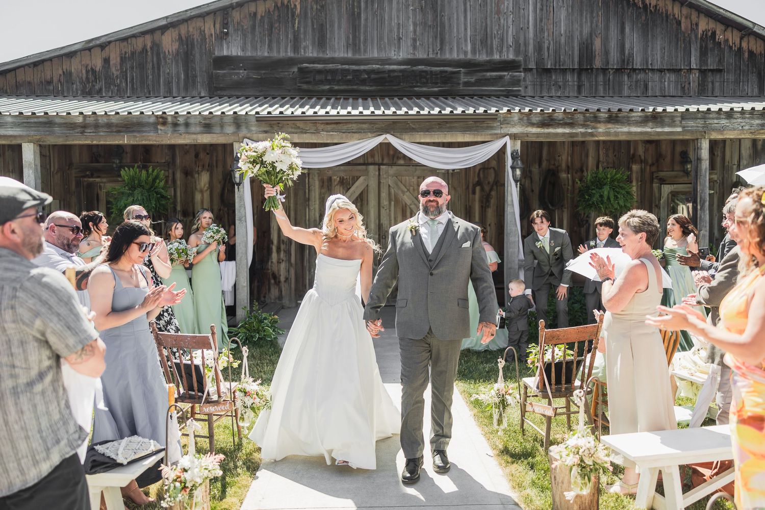 Newlyweds celebrate recessional as guests cheer outside rustic barn venue on sunny wedding day.