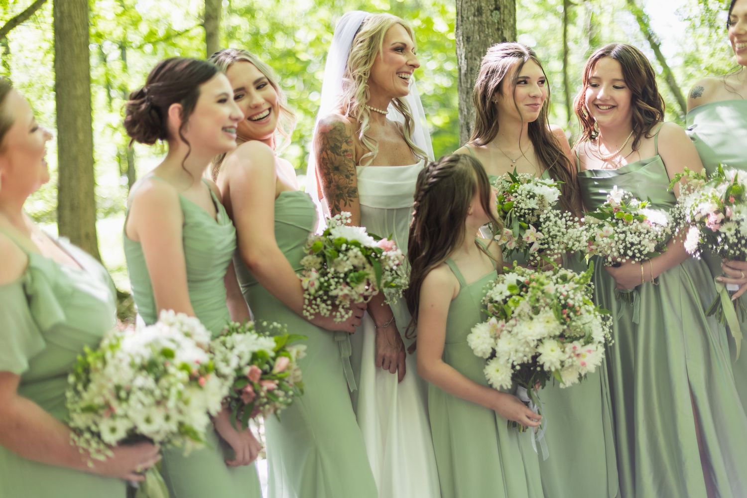 Bridesmaids in sage green dresses holding white floral bouquets laughing outdoors in a wooded setting.