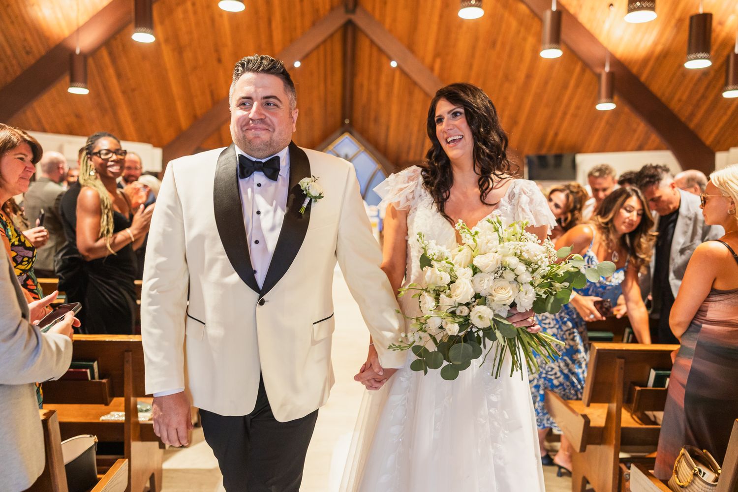 Bride and groom walk down the aisle in a wood-paneled church, she holds a white floral bouquet.