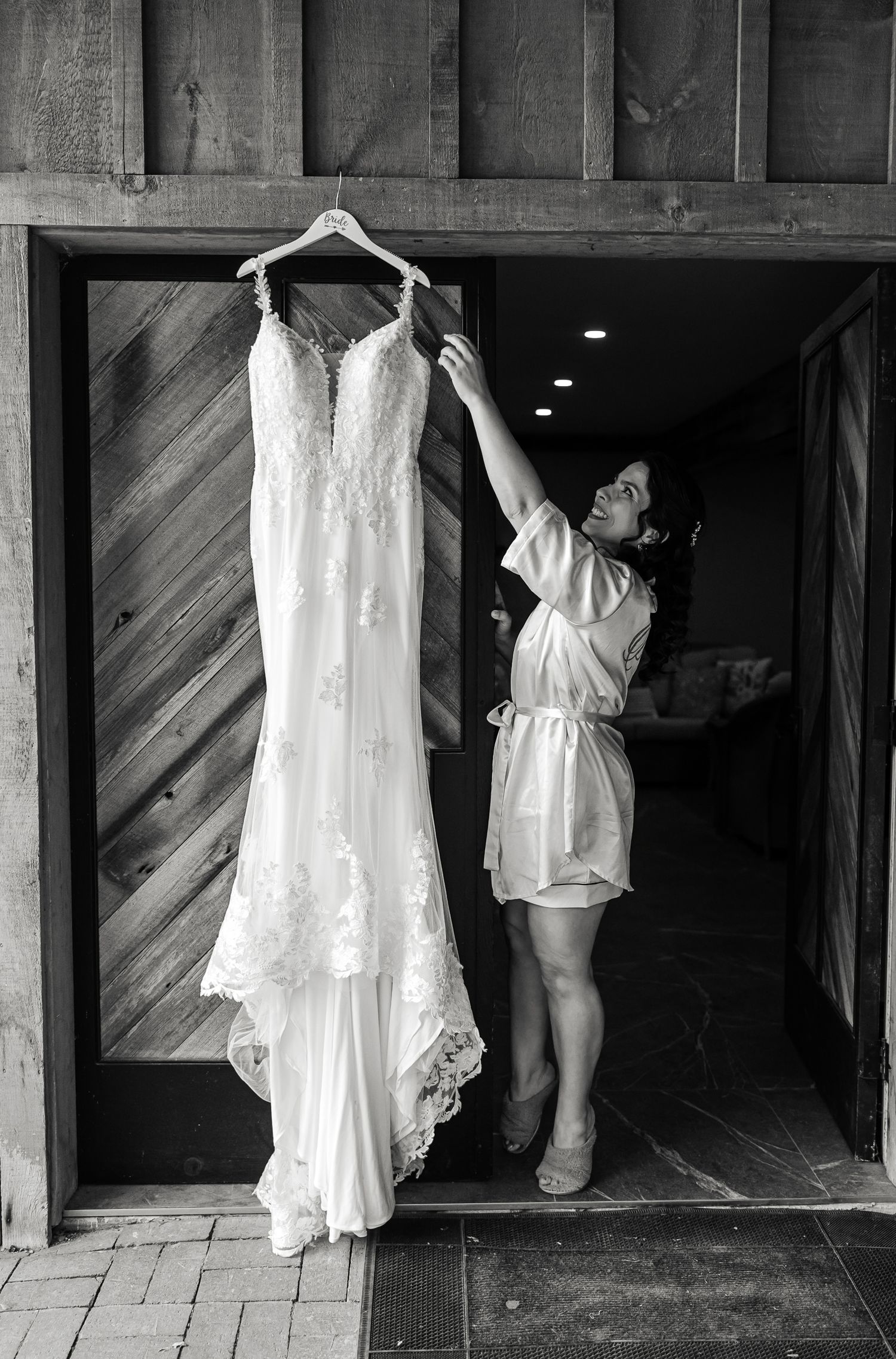 Bride in white robe adjusting her lace wedding gown hanging in a rustic barn doorway, black and white photo.