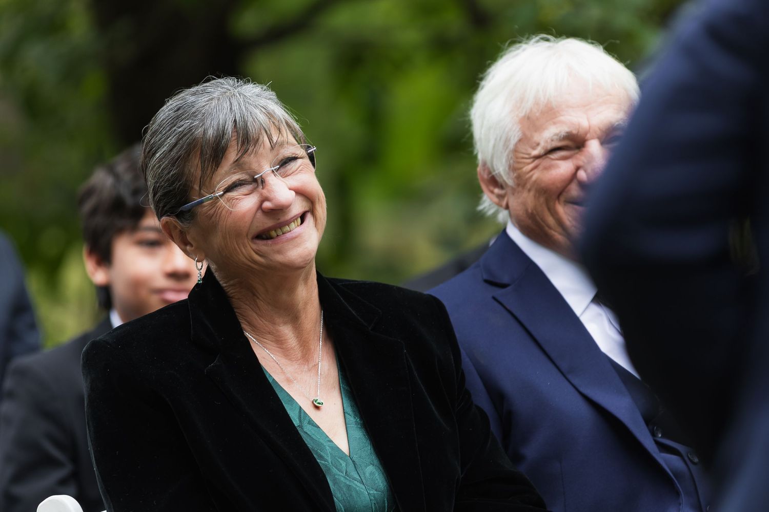 Elderly couple smiling joyfully outdoors at a wedding ceremony surrounded by greenery.