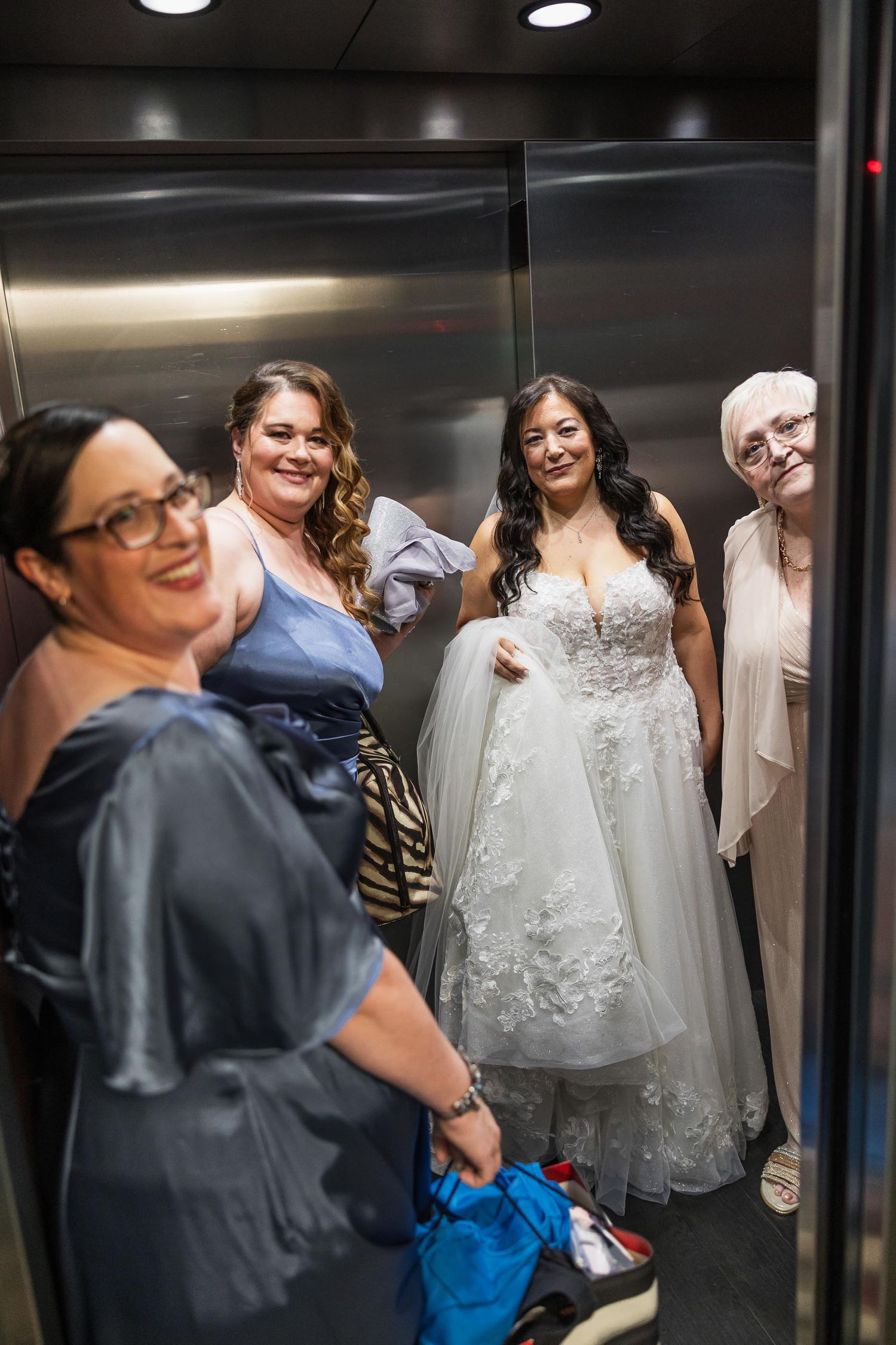A group of people gathered in an elevator with a bride wearing a white lace wedding gown.