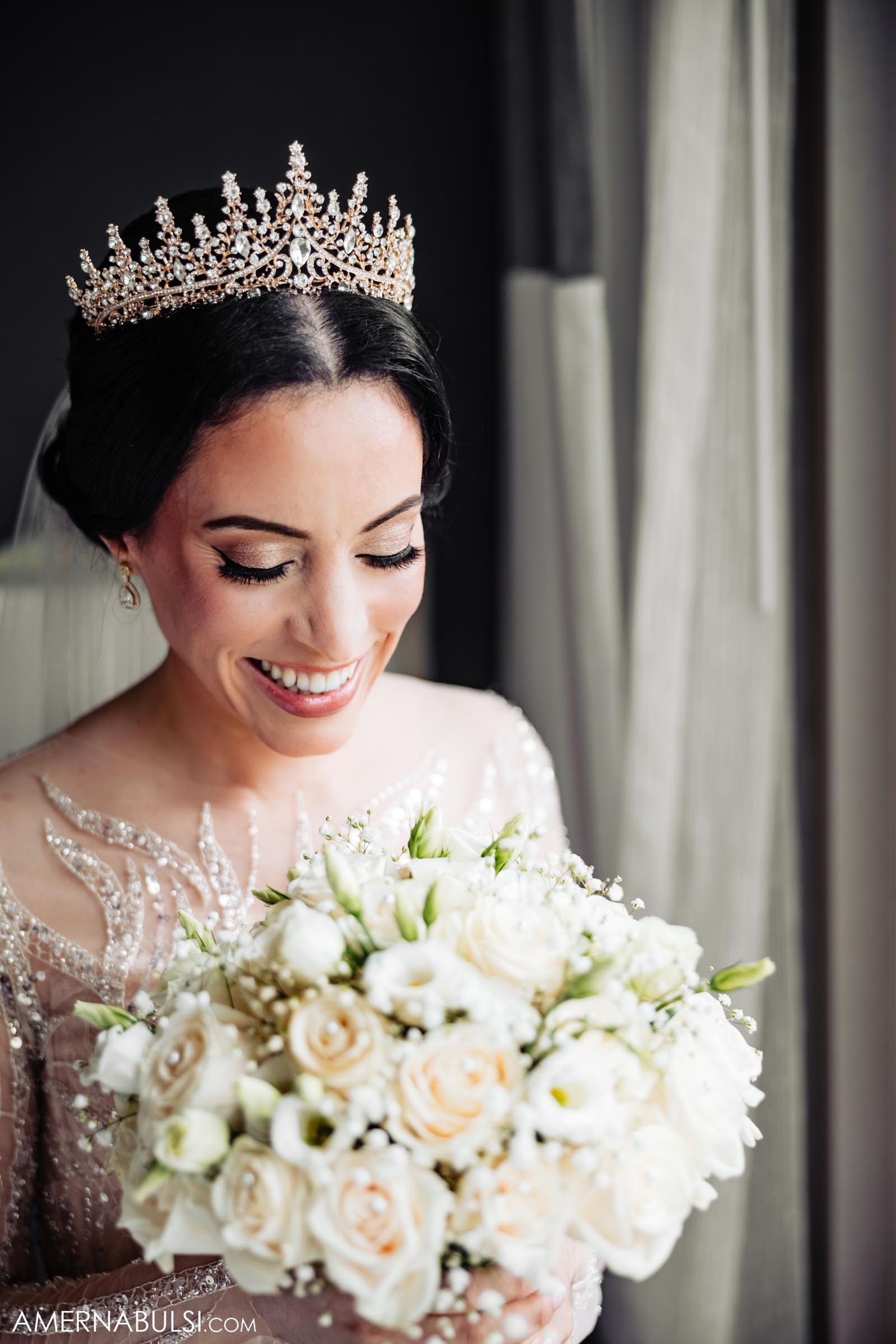 Close-up of bride wearing crystal crown and holding white rose bouquet while smiling.