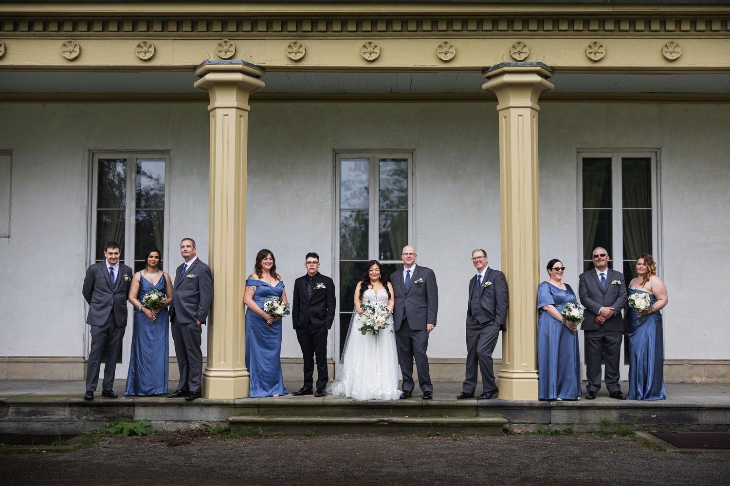 Wedding party in blue and gray attire posed formally on mansion's front steps.