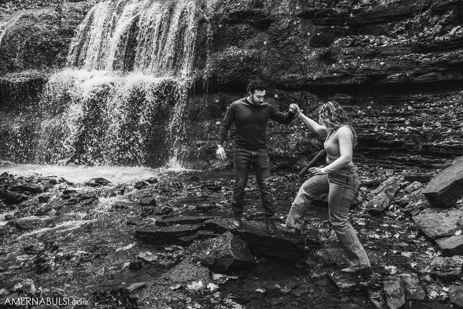 A couple explores the rocky base of a waterfall together in black and white.