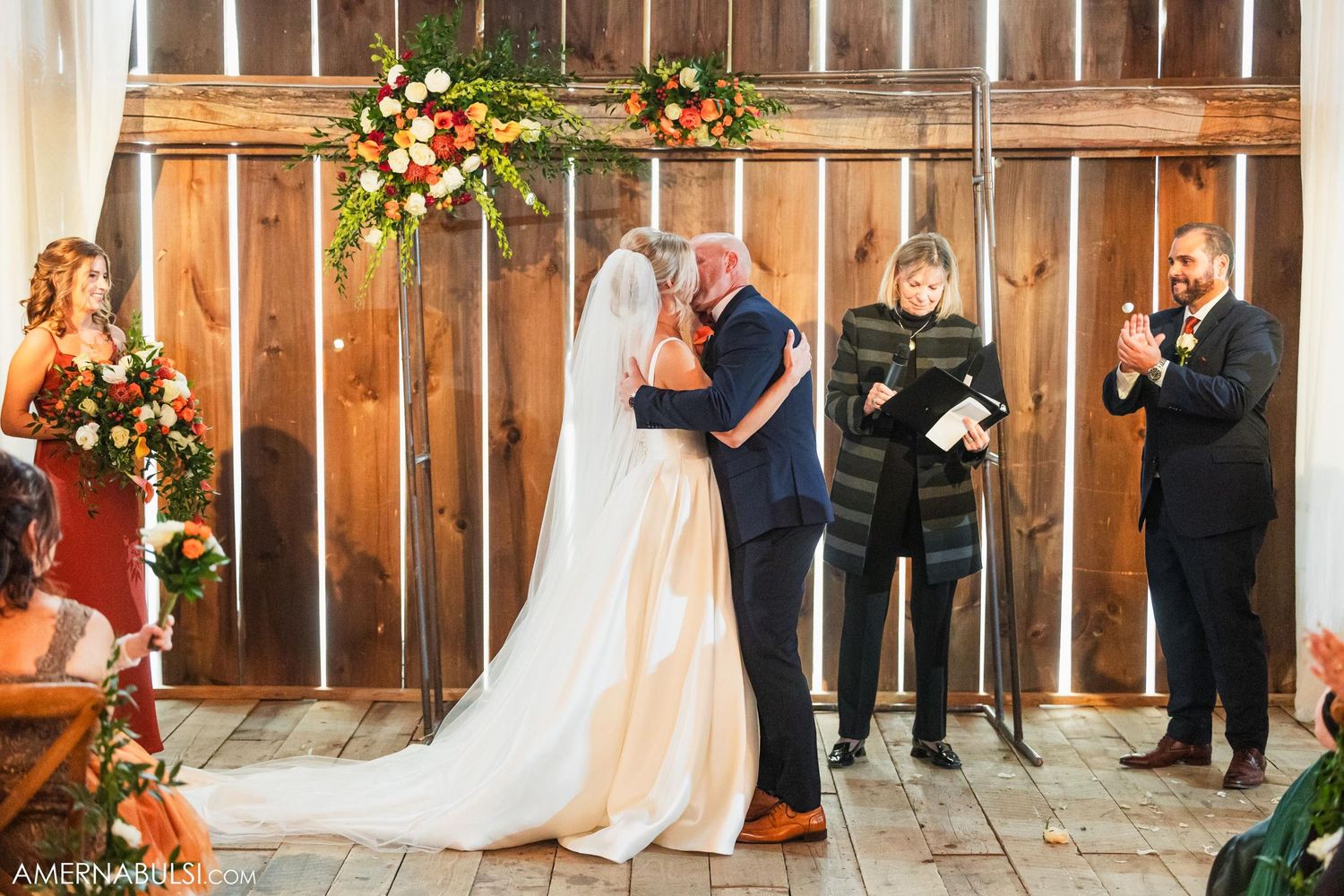 Wedding ceremony in a rustic barn setting with floral arch and guests looking on.
