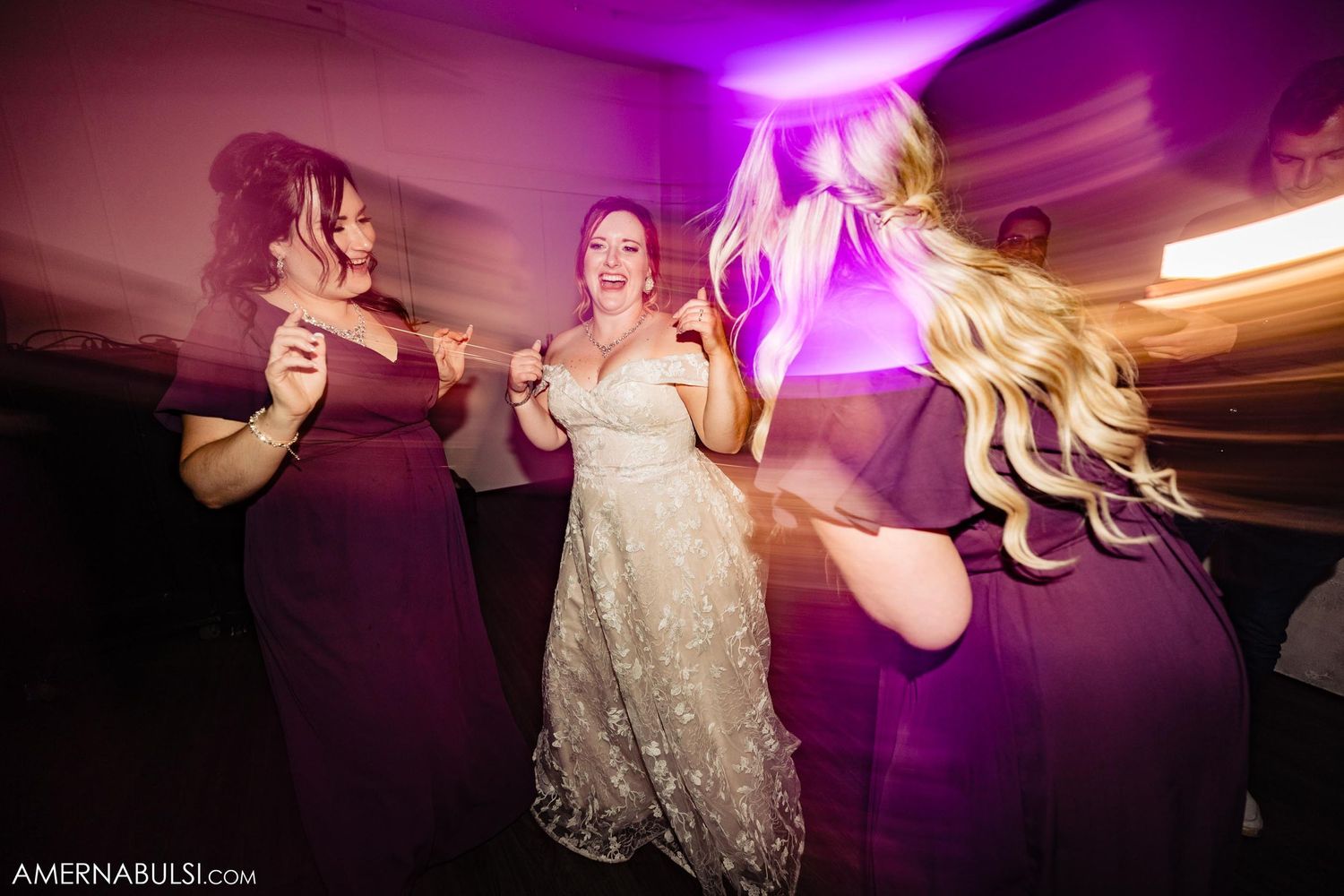 Group of wedding guests dancing enthusiastically under purple venue lighting.