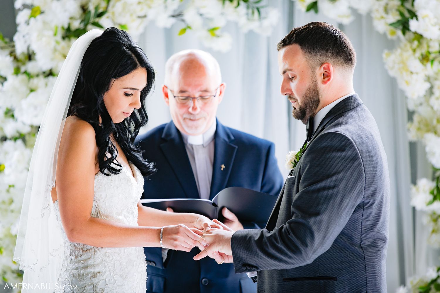 Wedding ceremony exchange of rings with officiant and floral arch backdrop.
