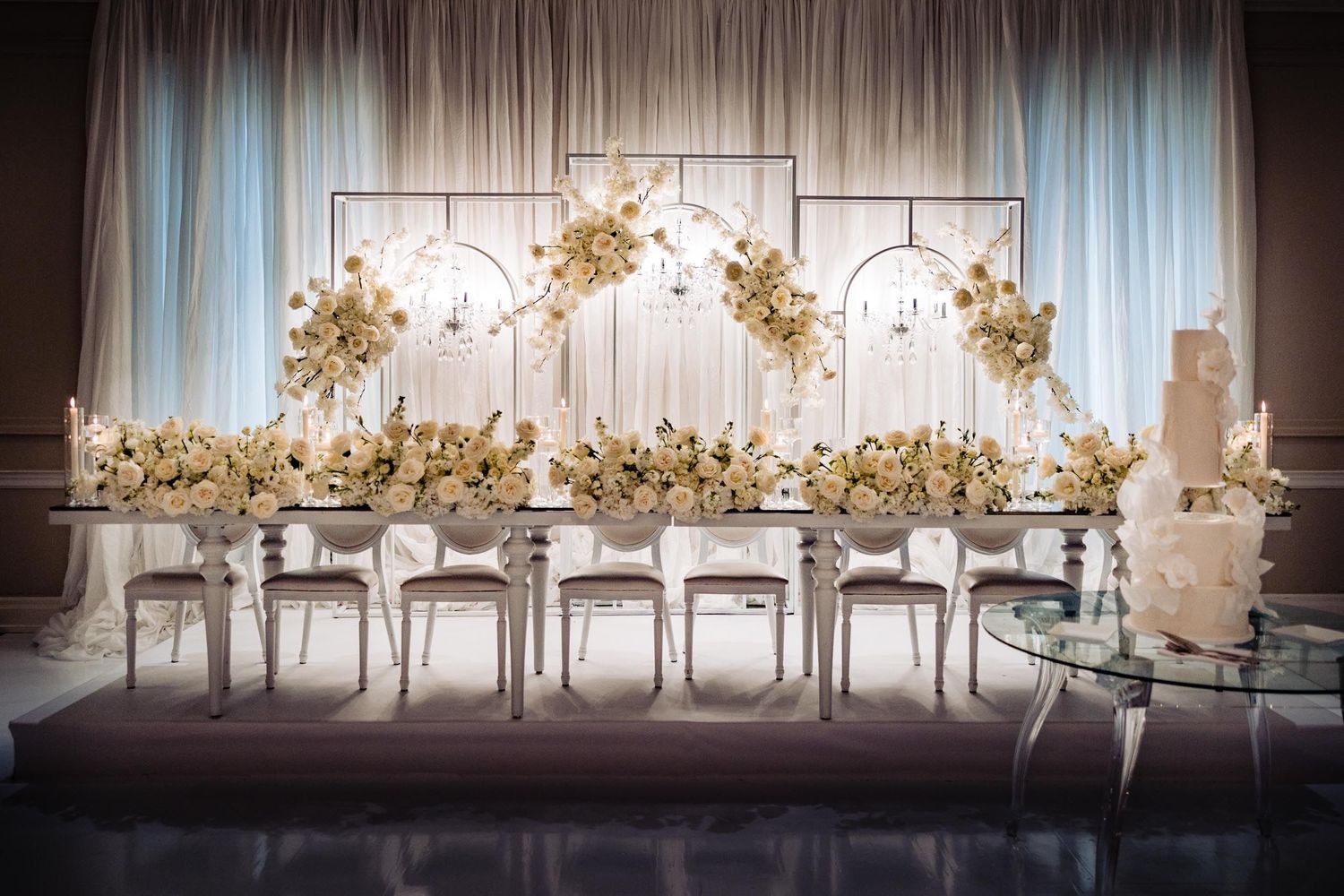White floral arch wedding head table setup with elegant chairs and decor.