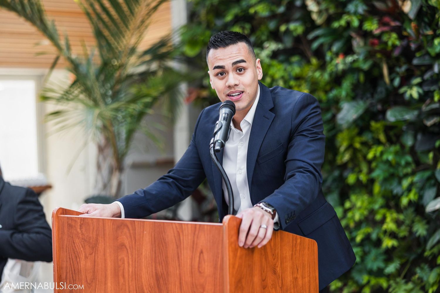 A speaker in a navy suit gives a speech at a podium during an outdoor event.