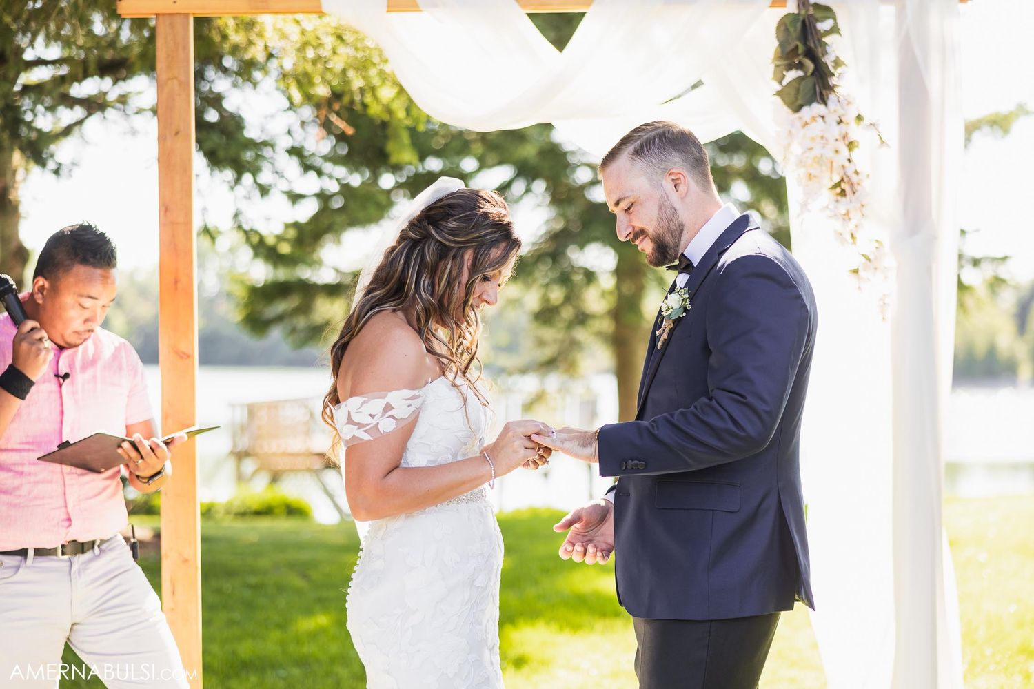 A couple exchanges rings during their outdoor wedding ceremony under a decorated wooden pergola.