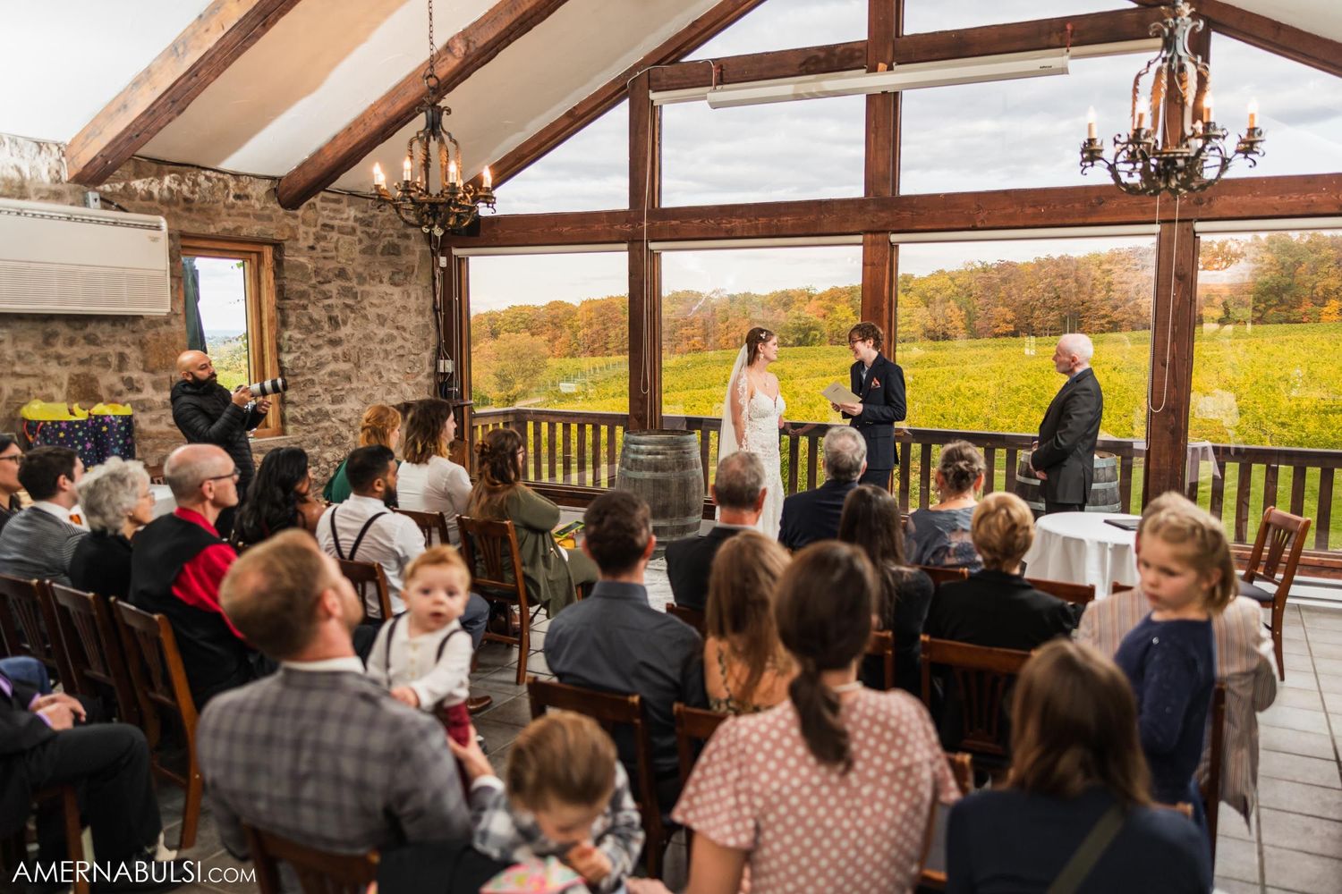 Wide view of an indoor wedding ceremony with guests seated facing large windows overlooking autumn landscape.