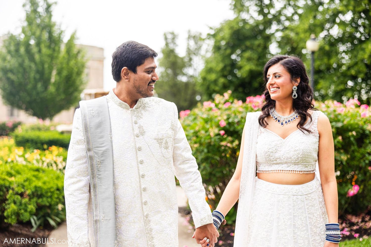 Couple in white Indian wedding attire walking together in a garden setting.