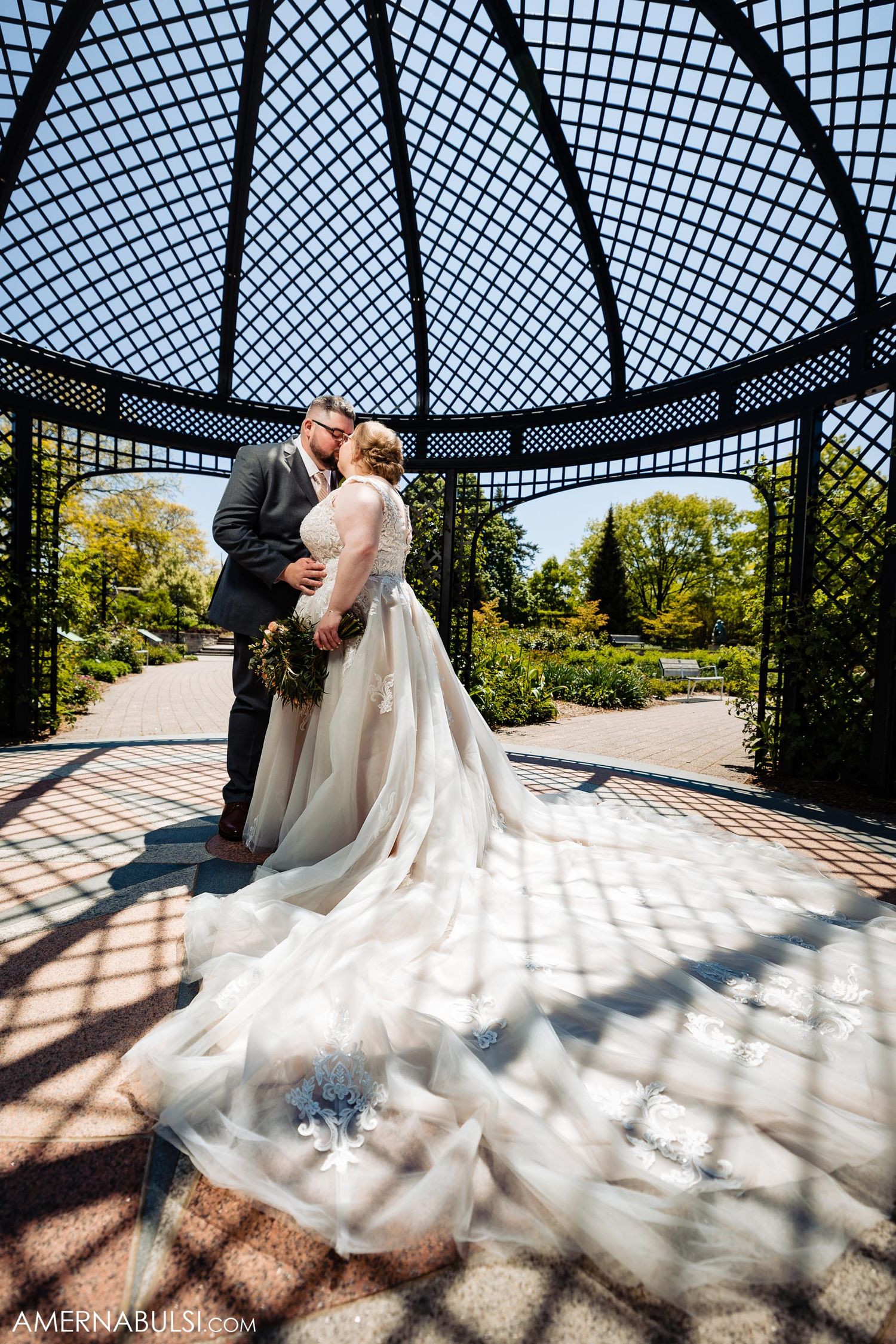 Wedding couple embraces in geometric dome gazebo with flowing white dress train.