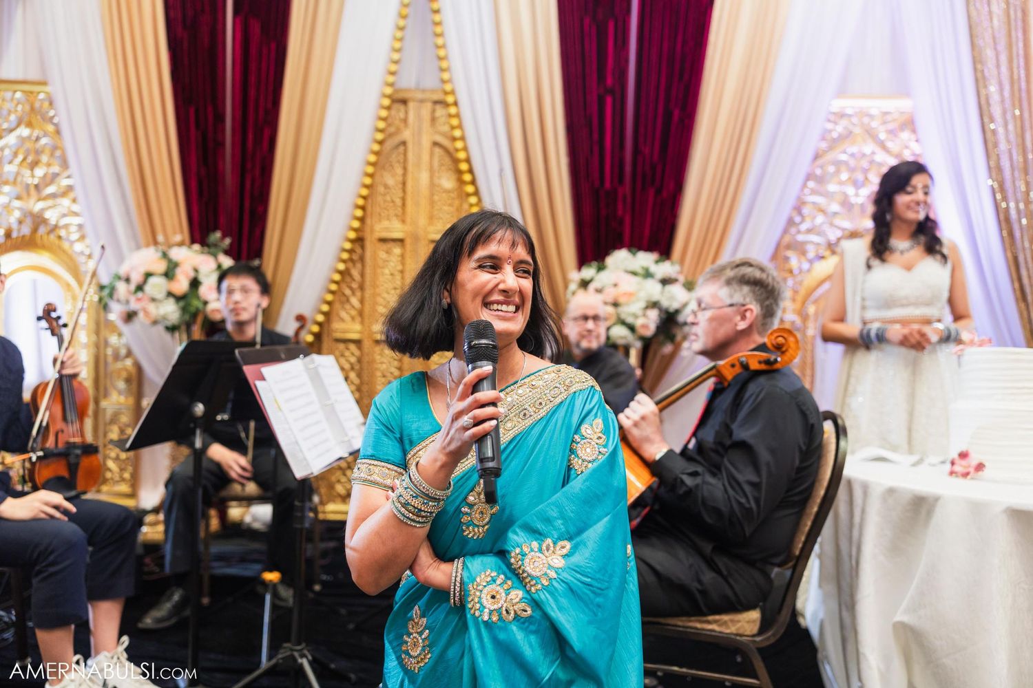 Guest in turquoise sari gives speech at wedding reception with live band in background.