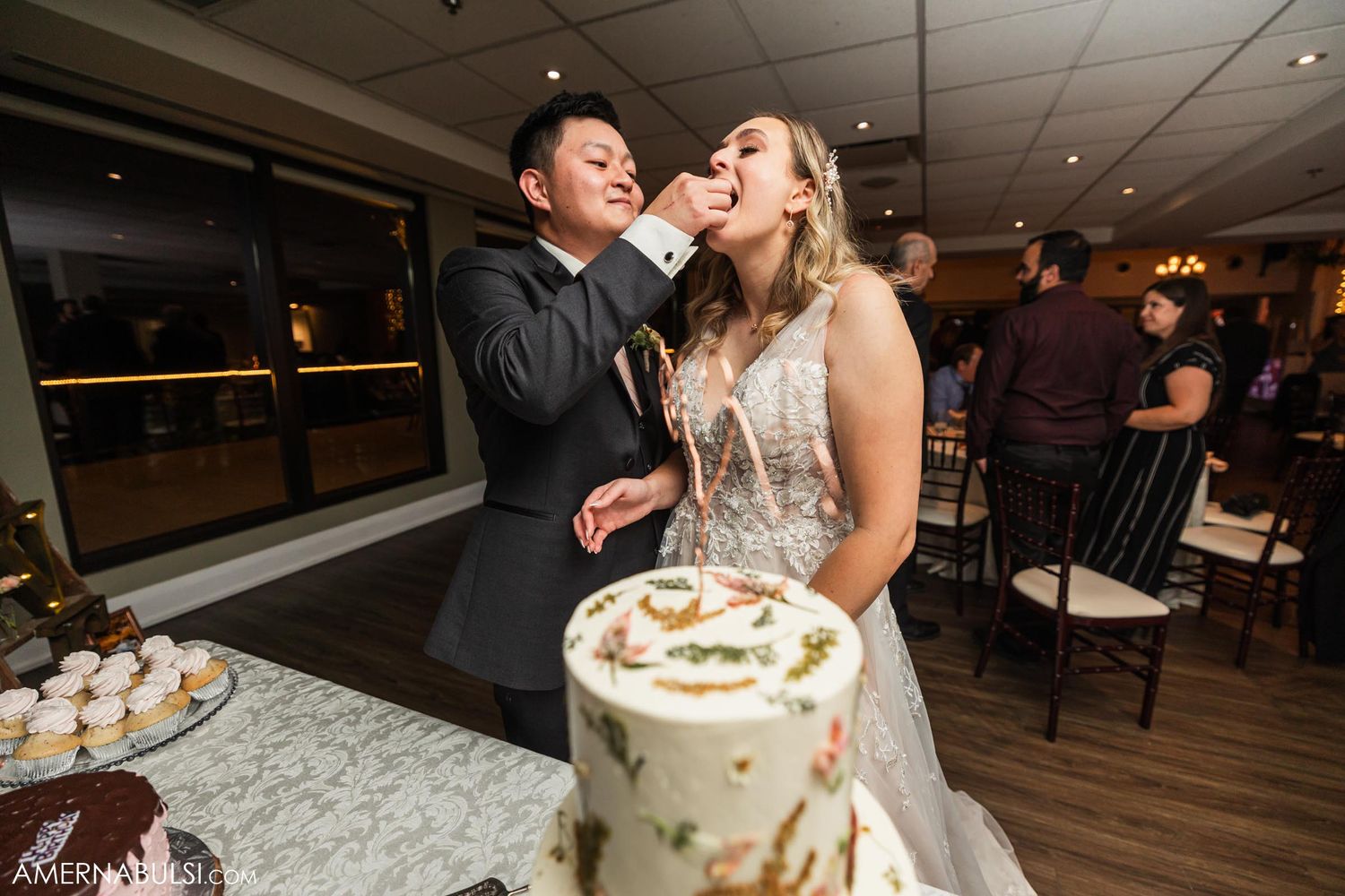 Newlyweds laugh while feeding each other wedding cake at their reception.