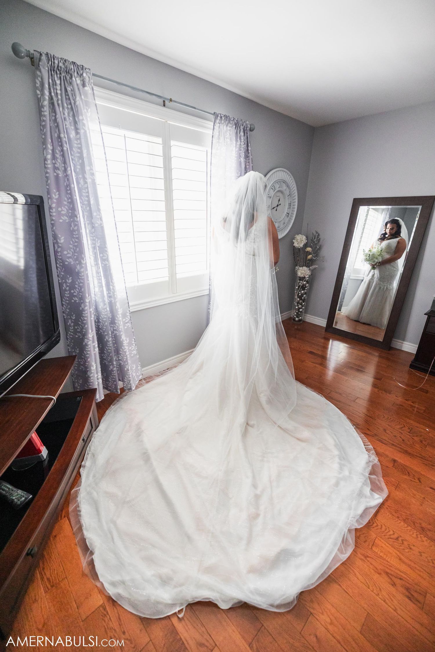 Long flowing wedding dress train and veil displayed in room with mirror and gray curtains.