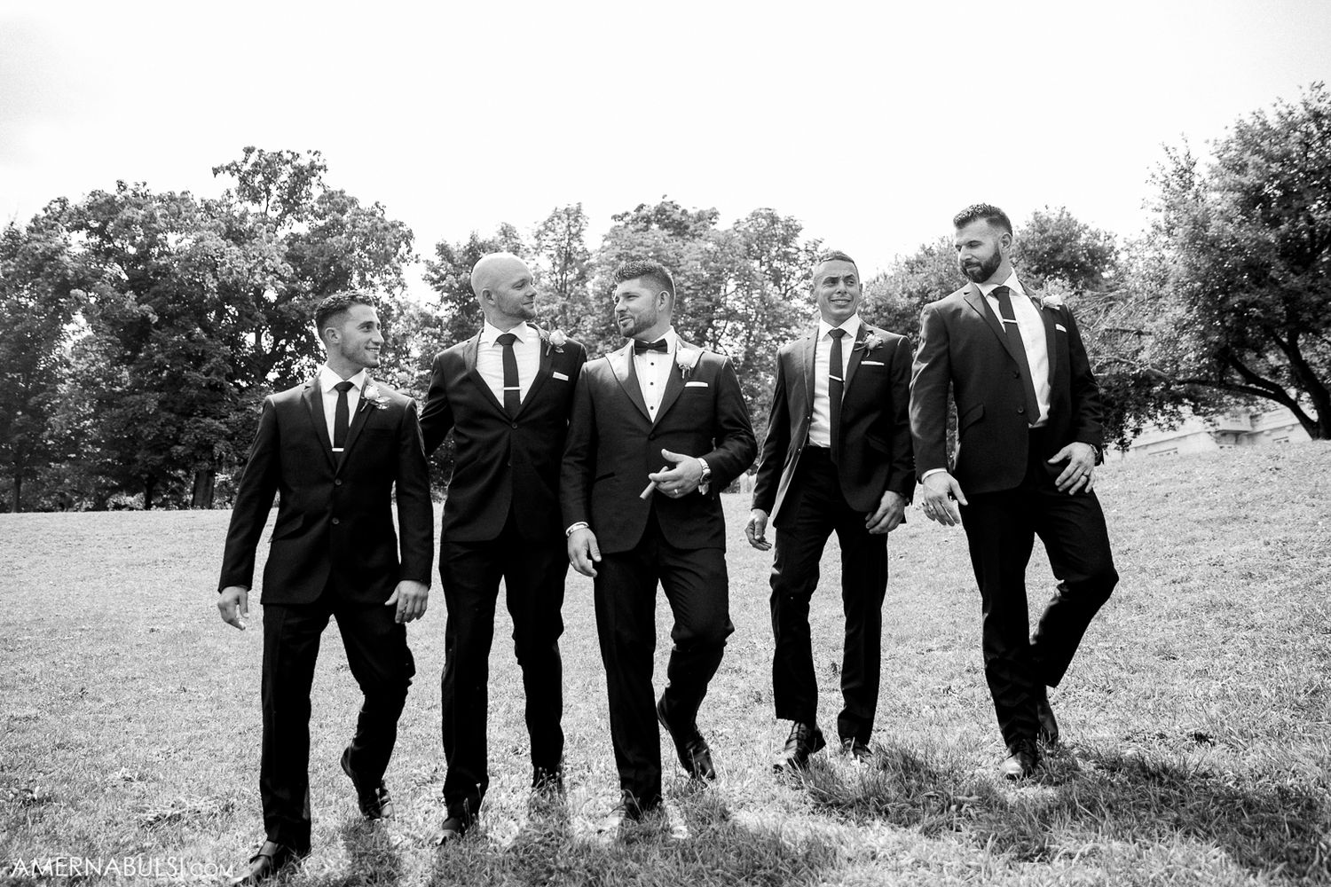 Groomsmen in black suits pose together outdoors in a natural field setting.