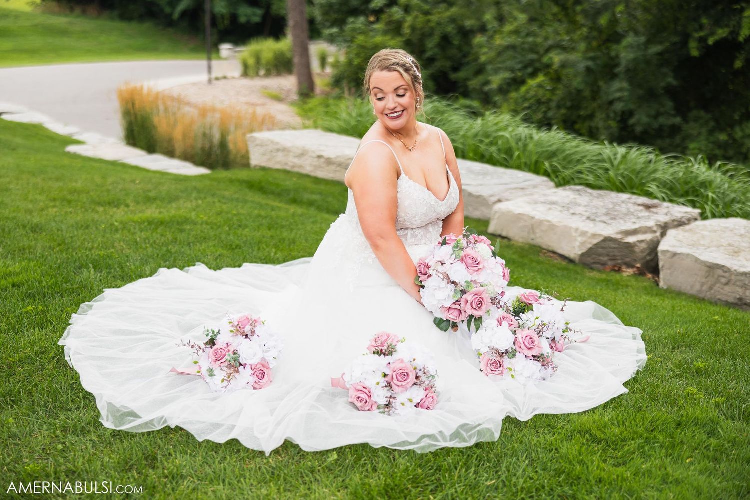 Bride in white lace gown with floral details poses seated on grass.