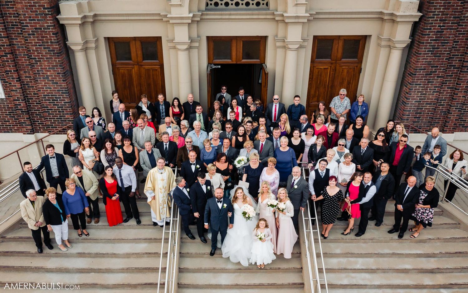 Large wedding party gathers on classic brick building steps for formal group portrait.