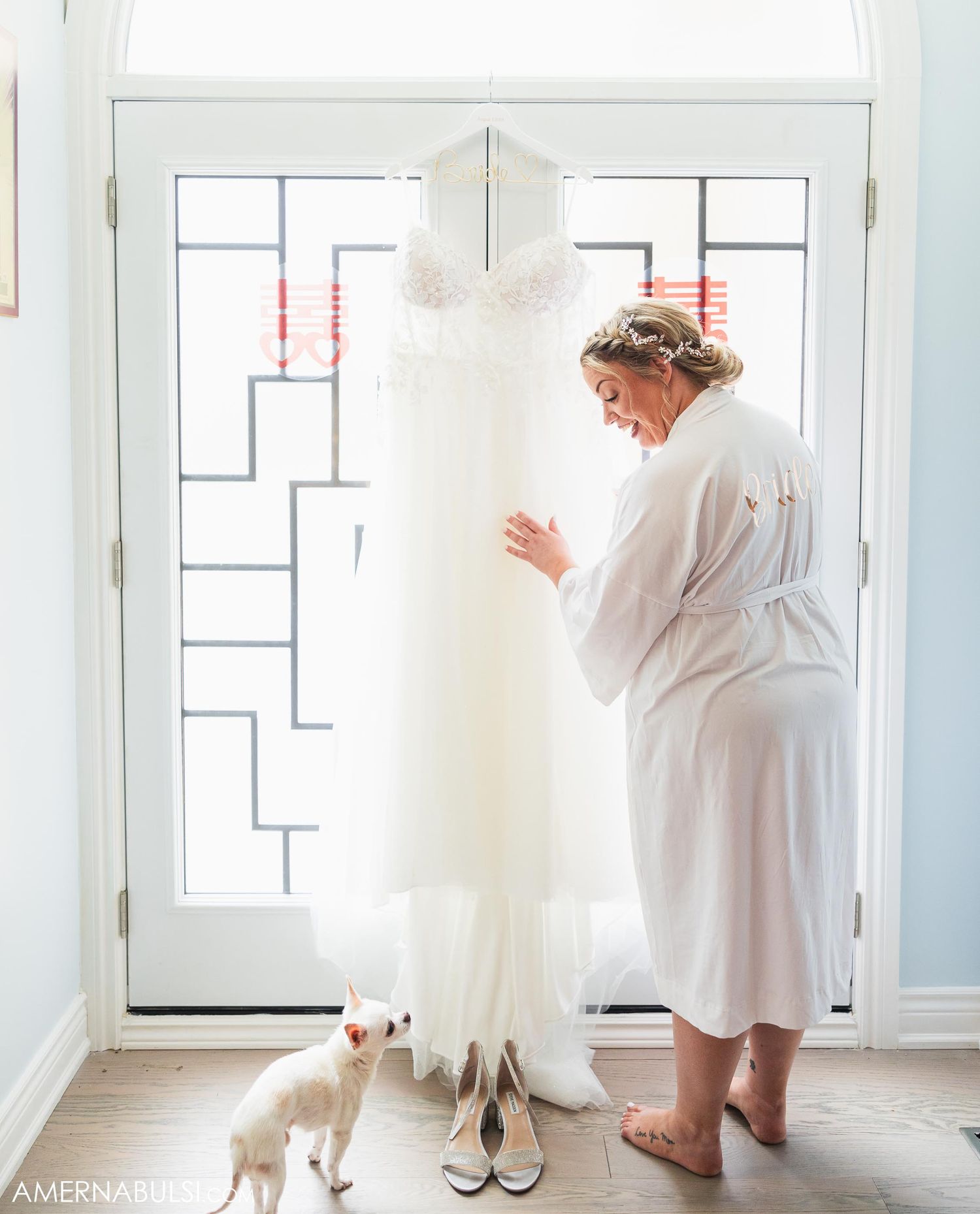 White wedding dress hanging by window while person in robe examines it with small dog nearby.