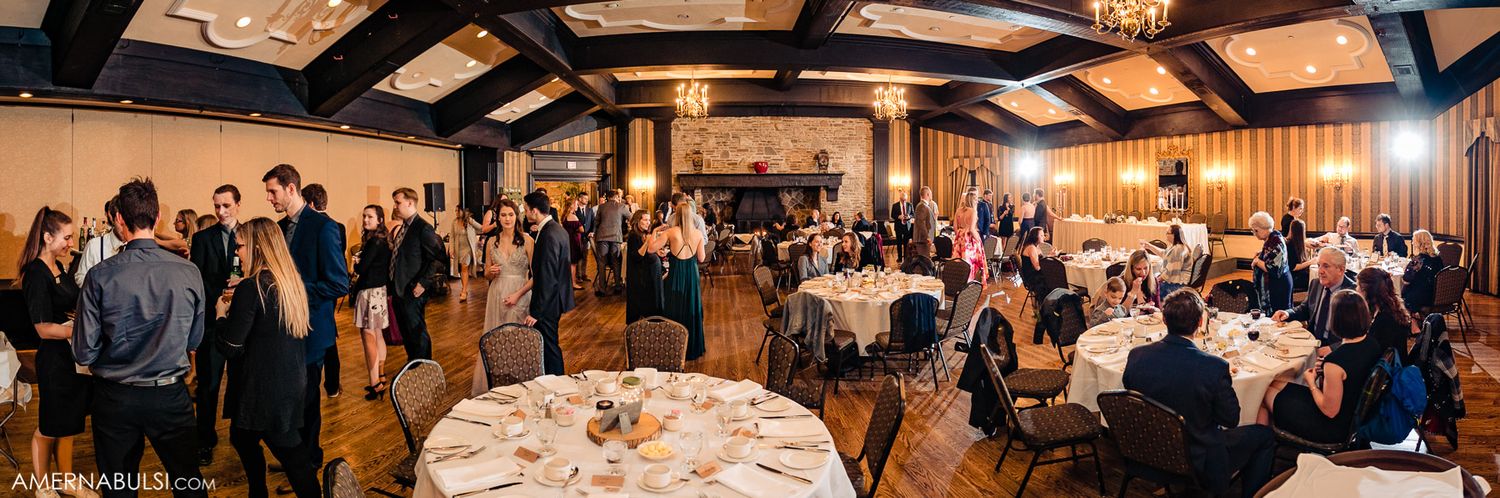 Panoramic view of a wedding reception hall with guests seated at decorated tables under warm lighting.
