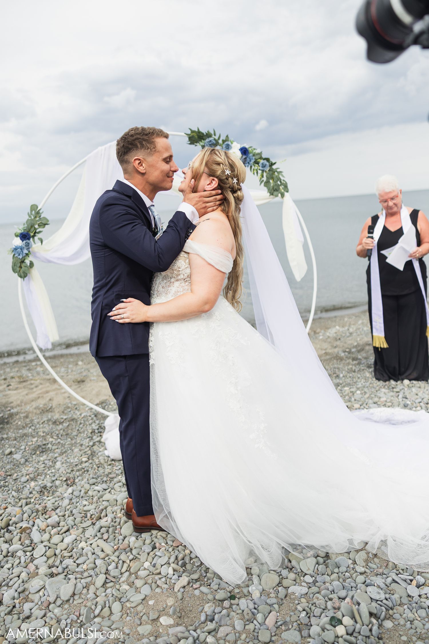 Bride and groom share a romantic moment with flowing veil on a rocky beach with cloudy skies in the background.