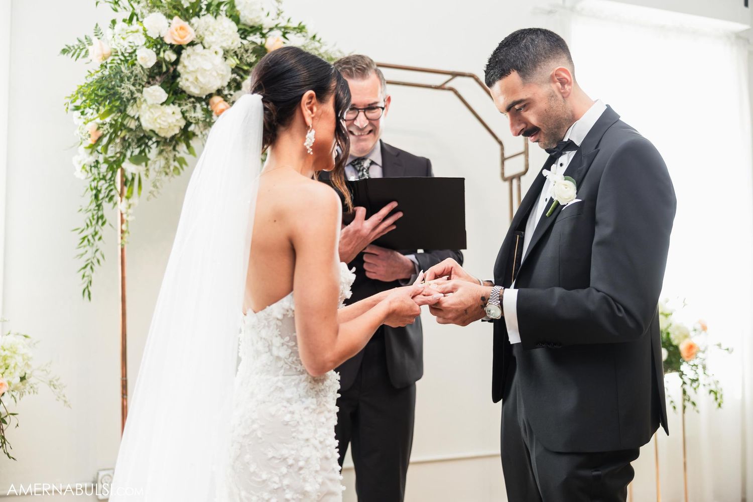 Wedding ceremony under a floral arch with couple exchanging rings as officiant holds ceremony book.