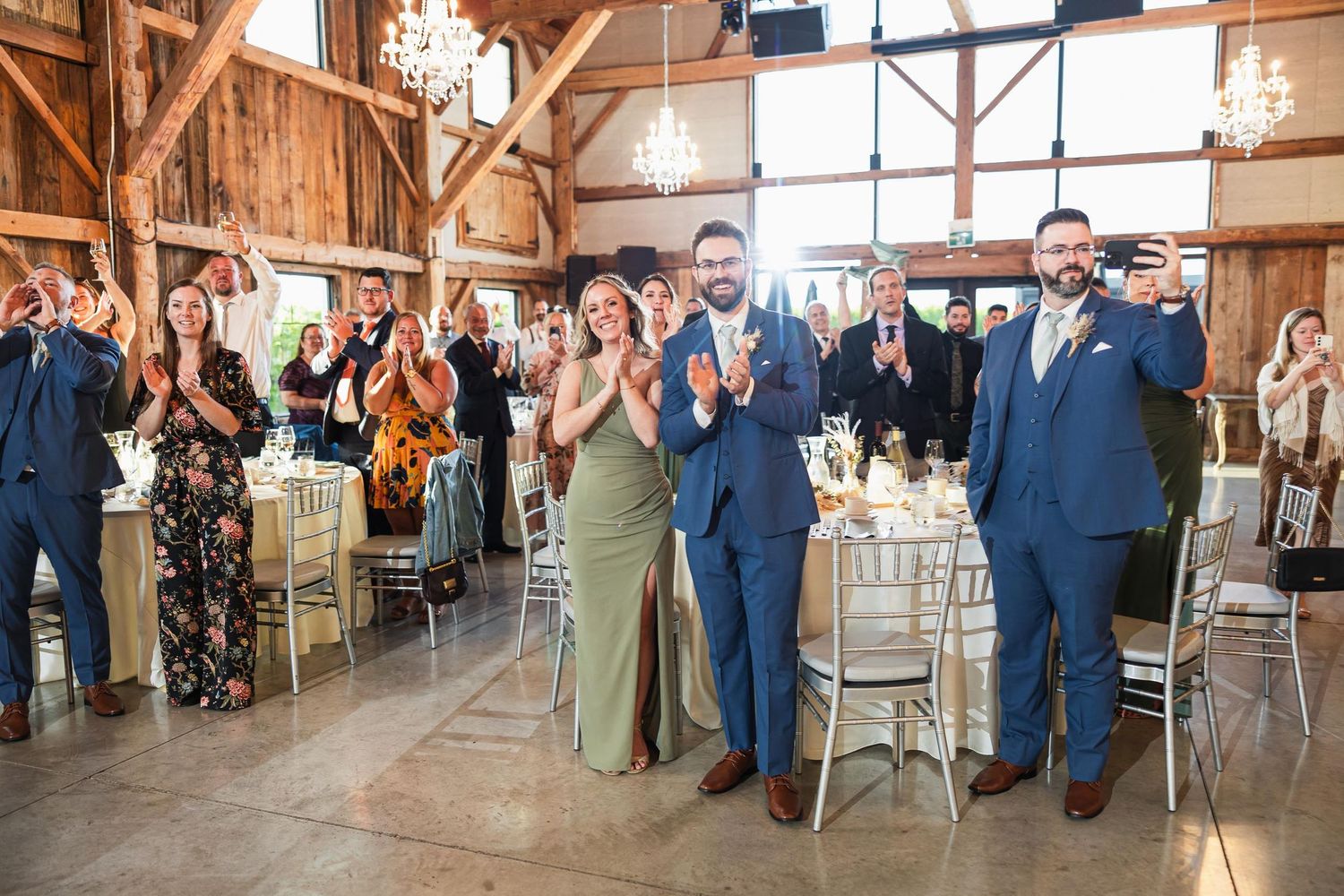 Wedding guests stand and applaud in a rustic barn venue with wooden beams and chandeliers.