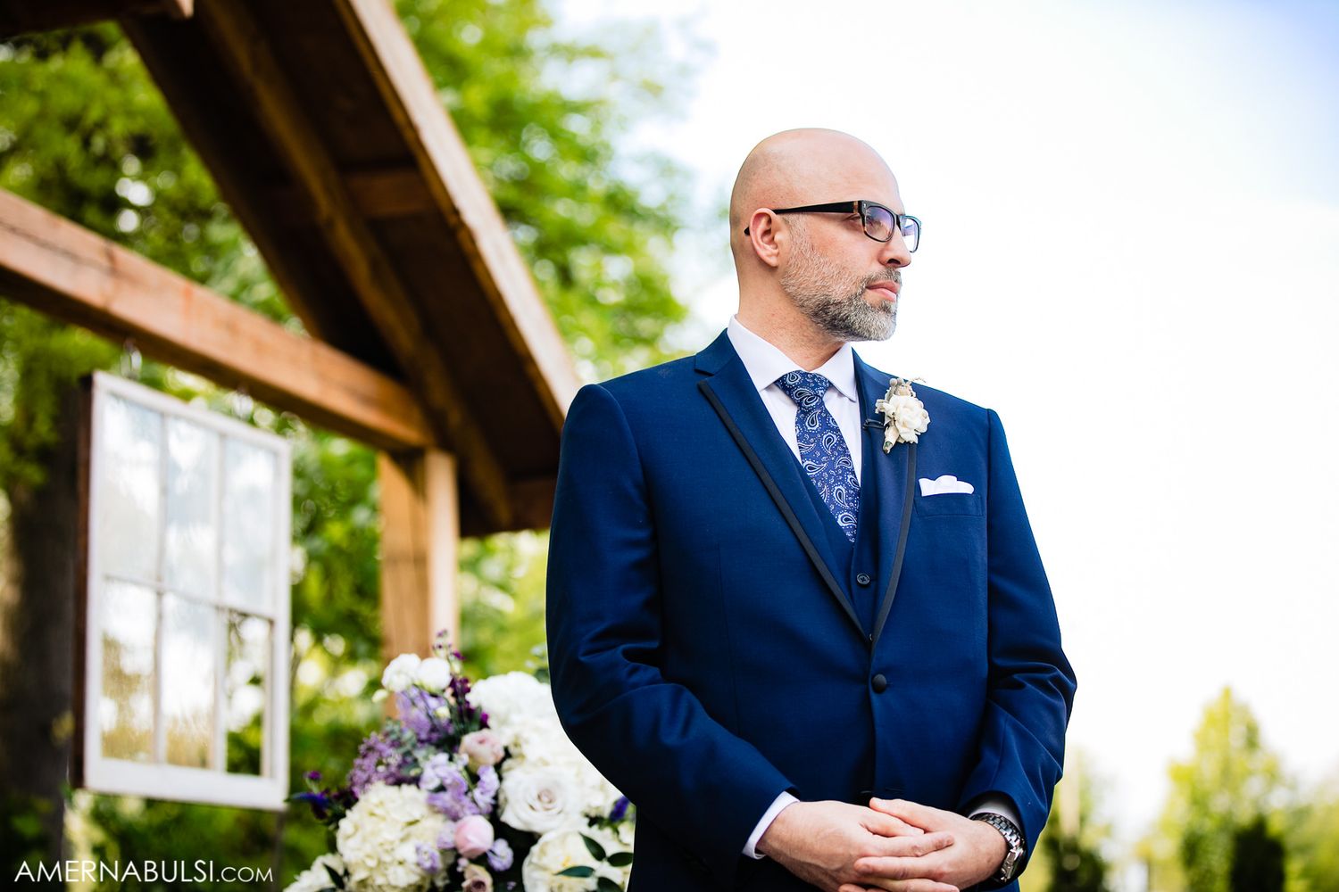 Wedding party members in navy suits and boutonnieres stand together at an outdoor ceremony.