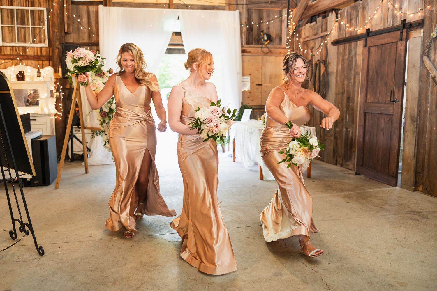 Wedding party members dance in a line formation inside a vintage barn with wooden walls.