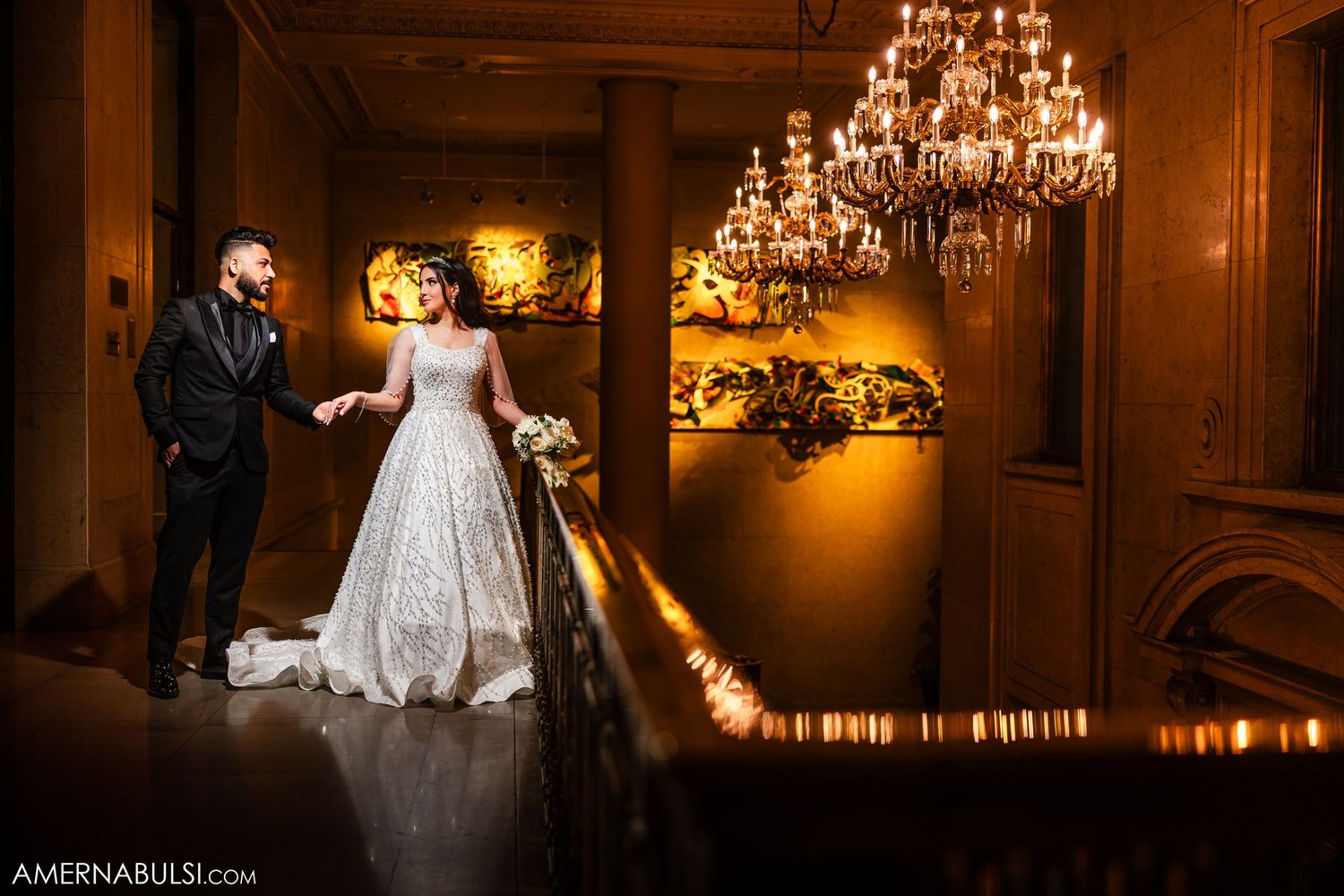 A bride and groom pose on a grand staircase beneath ornate chandeliers in a warmly lit venue.