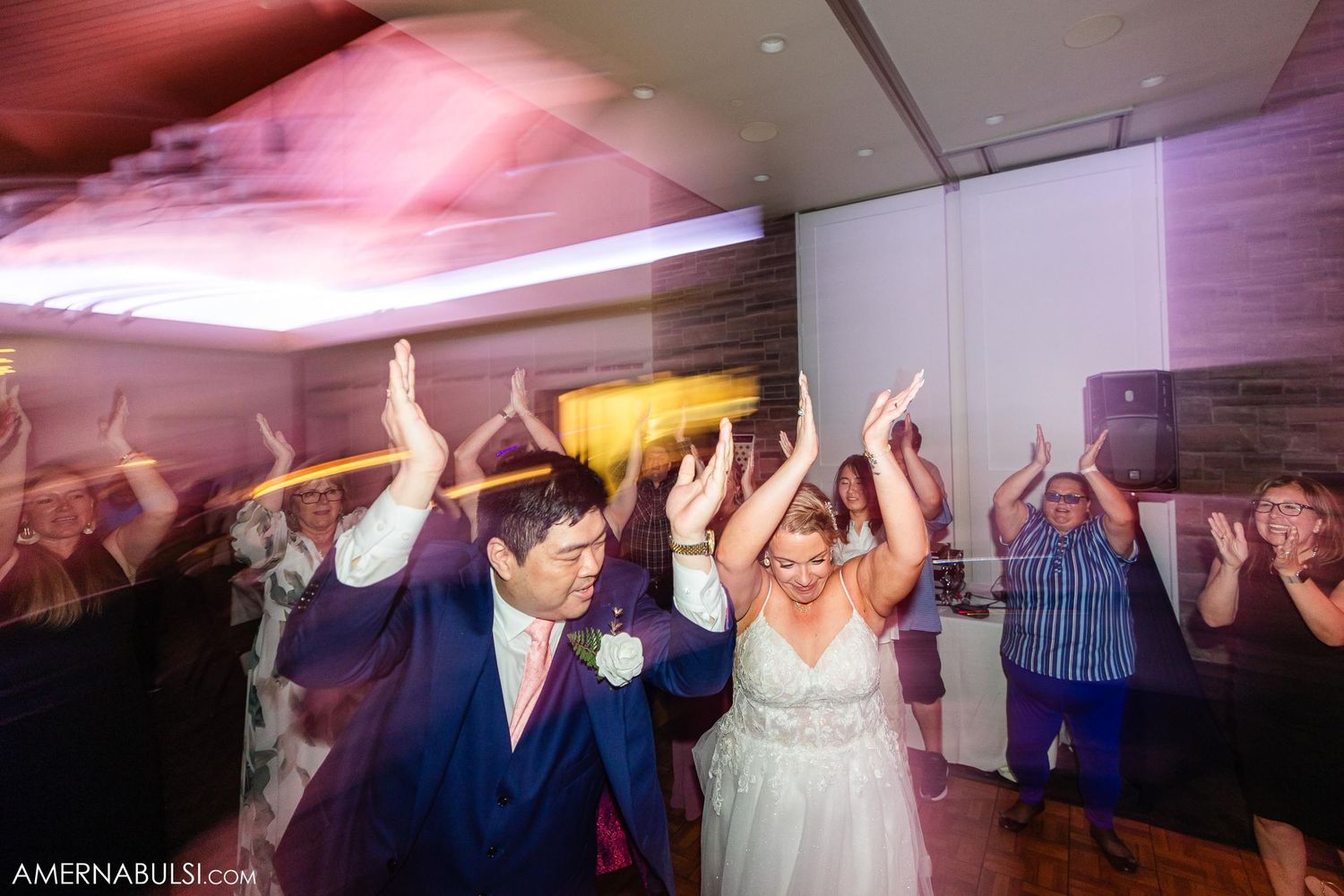 Energetic dance party at wedding reception with pink ceiling lighting.