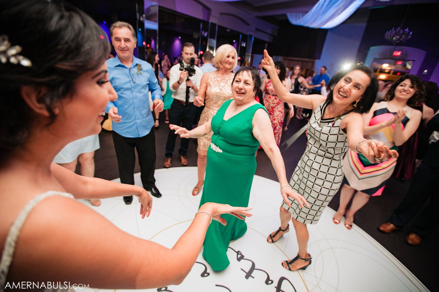 Wedding guests dance enthusiastically on a white dance floor at a reception with purple lighting.