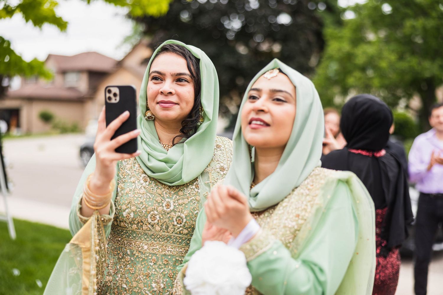 Two wedding guests in mint green hijabs smile while taking photos on their phones.