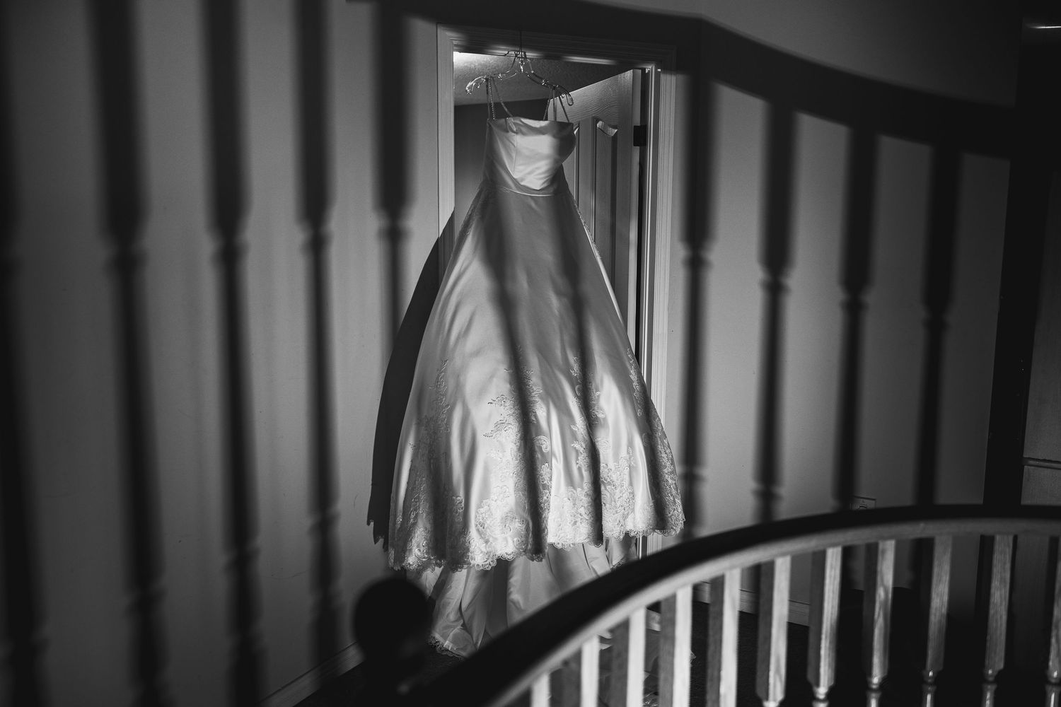 Black and white photo of a wedding dress hanging on staircase banister in elegant home interior.