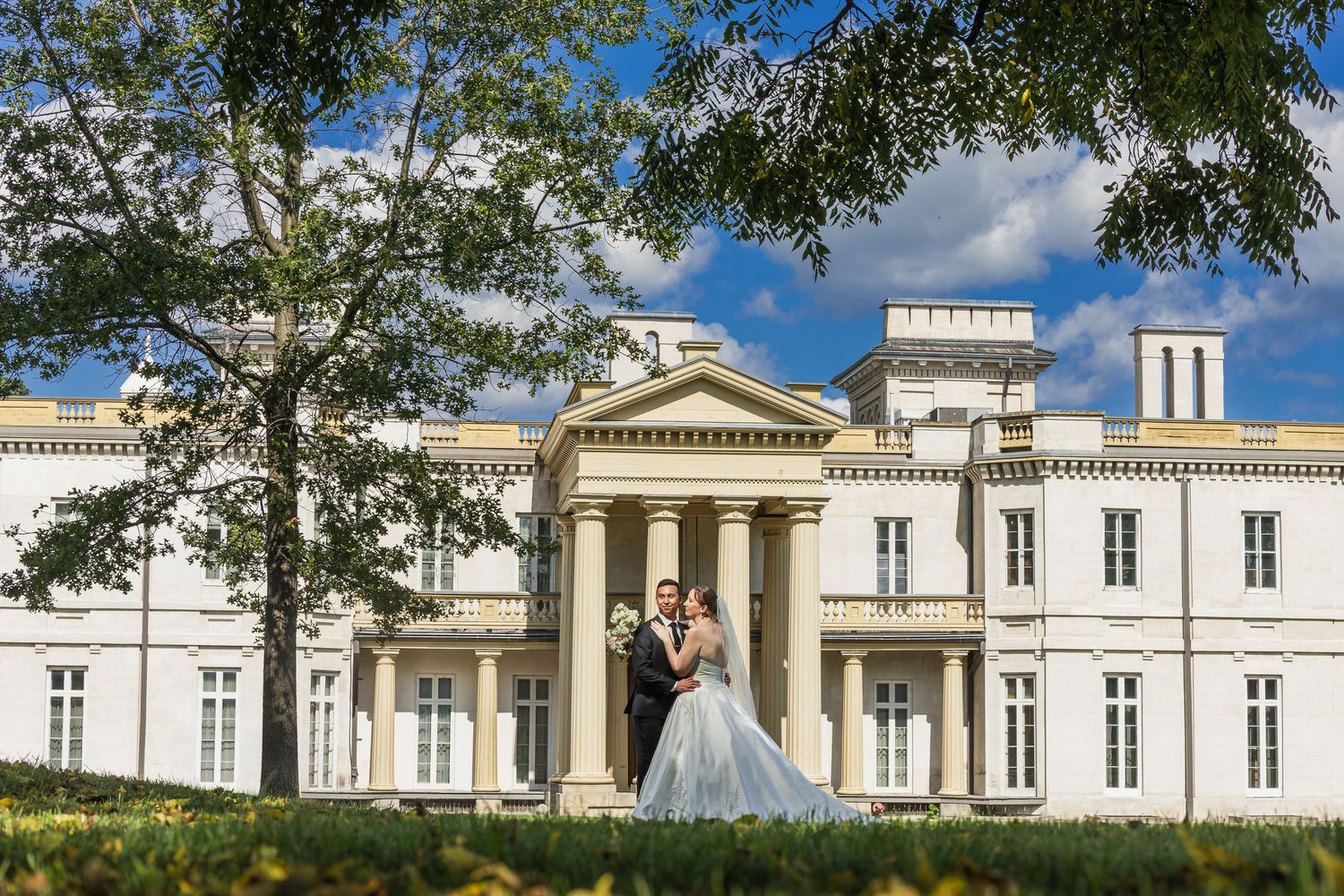 Bride and groom embrace in front of grand white neoclassical wedding venue.