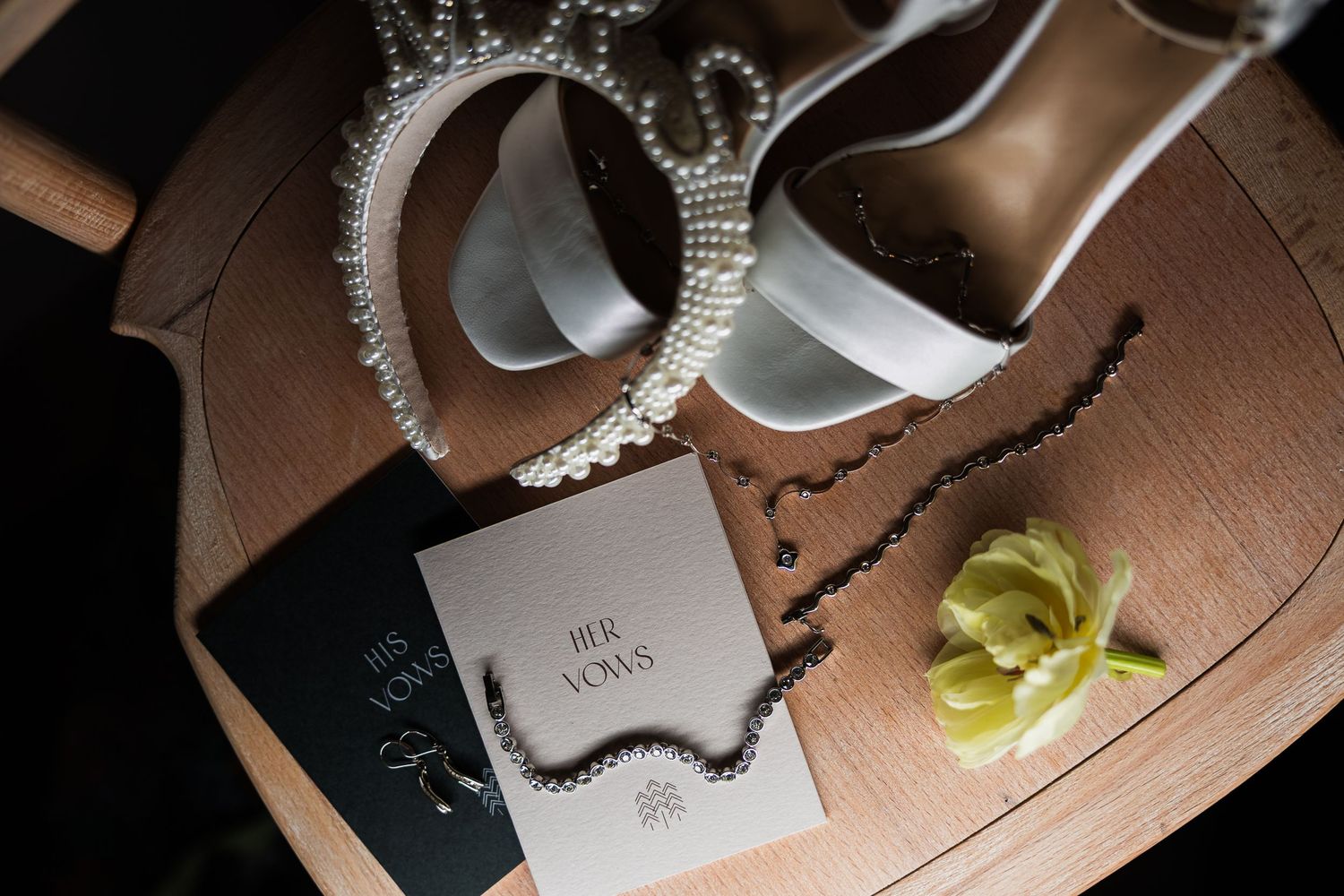 Wedding flat lay with pearl tiara, heels, vow booklets, rosary, and yellow flower on wooden surface.