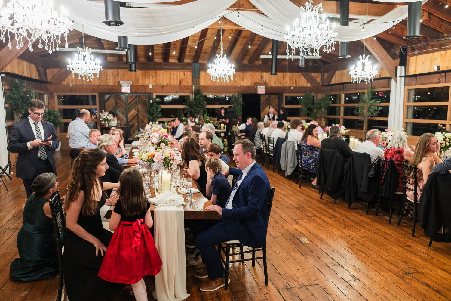 Elegant wedding reception in a rustic barn venue with chandeliers, draped ceilings, and guests seated at long decorated tables.