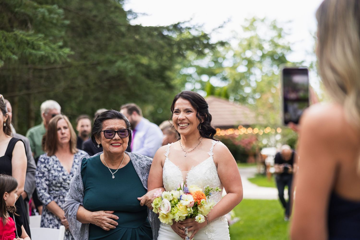 Smiling bride in white gown holding bouquet walks down aisle with family member at outdoor wedding.