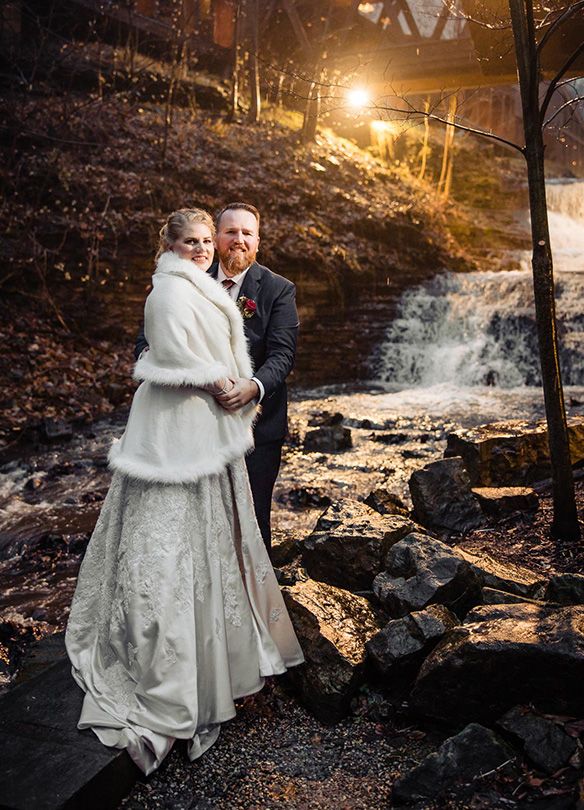 Couple embraces by illuminated waterfall at dusk in winter setting.