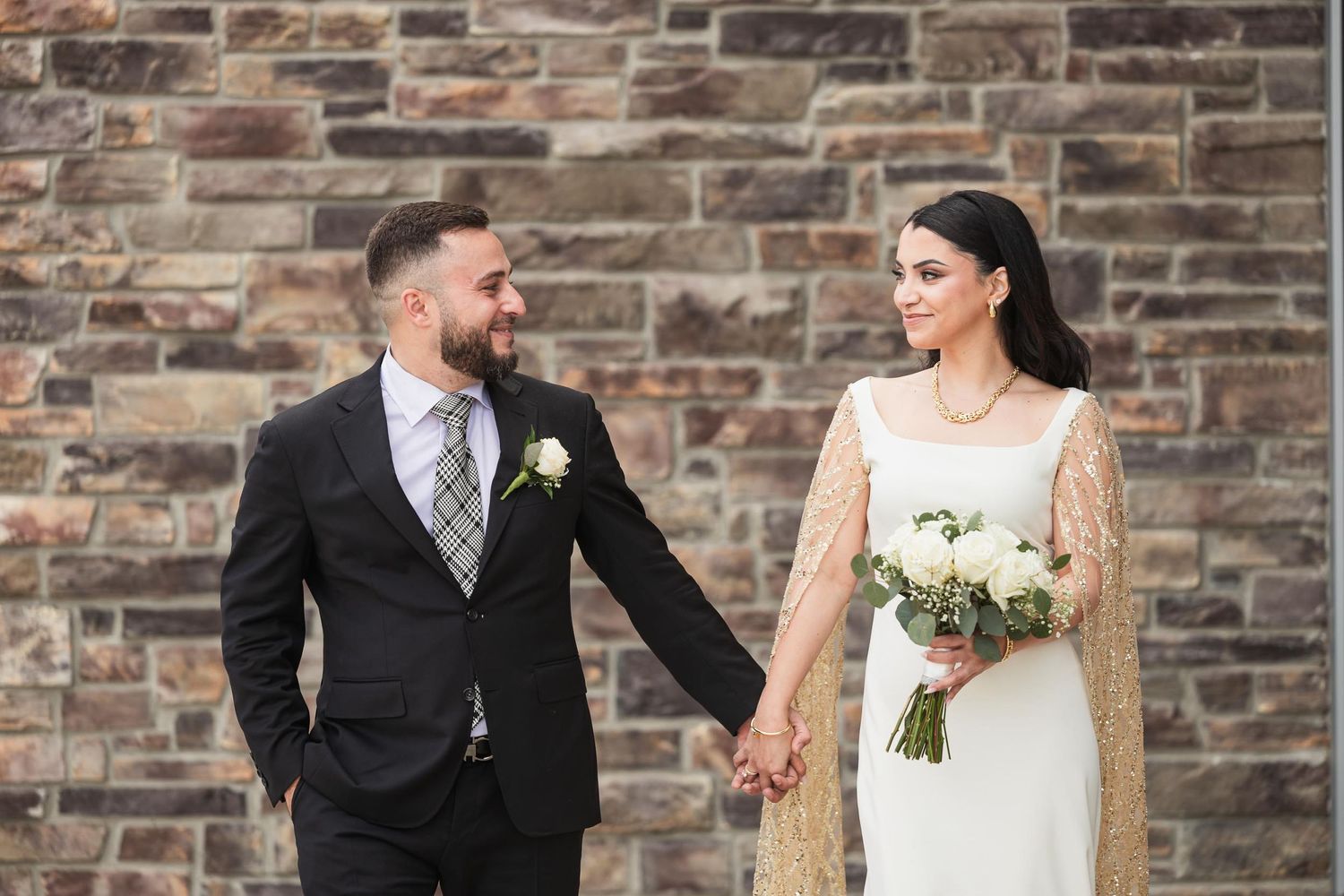 Couple in formal attire pose against rustic stone wall background.