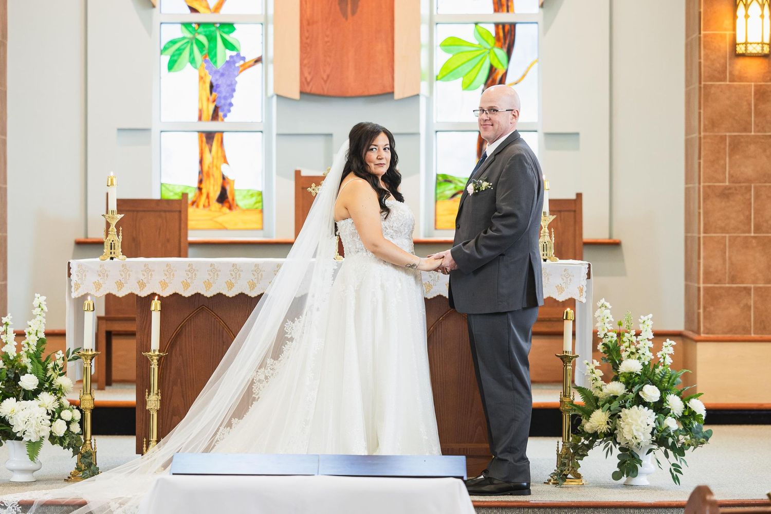 A traditional Catholic wedding ceremony takes place at the altar with floral decorations and stained glass windows in view.