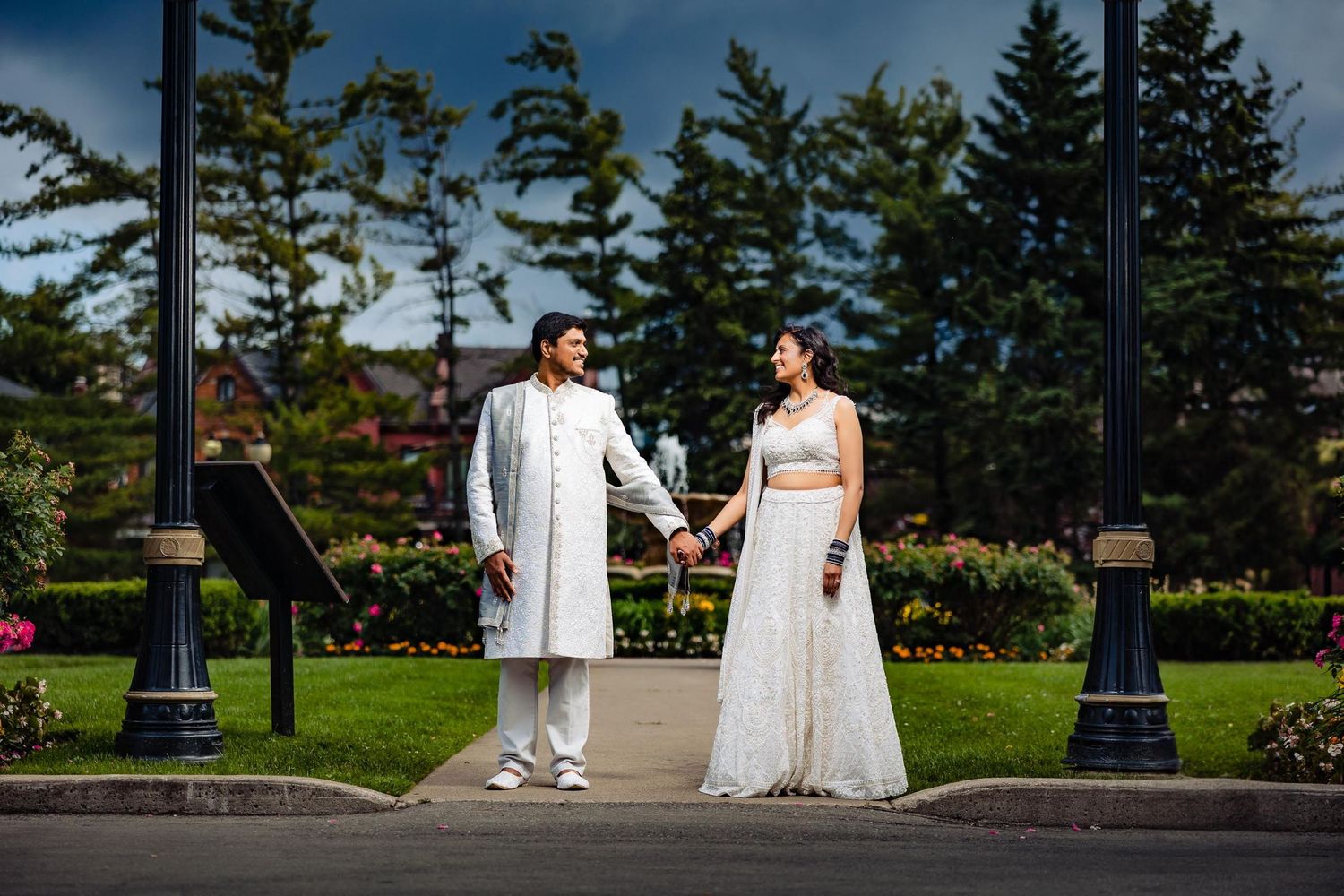 A couple stands together on a pathway with evergreen trees and lamp posts in the background.