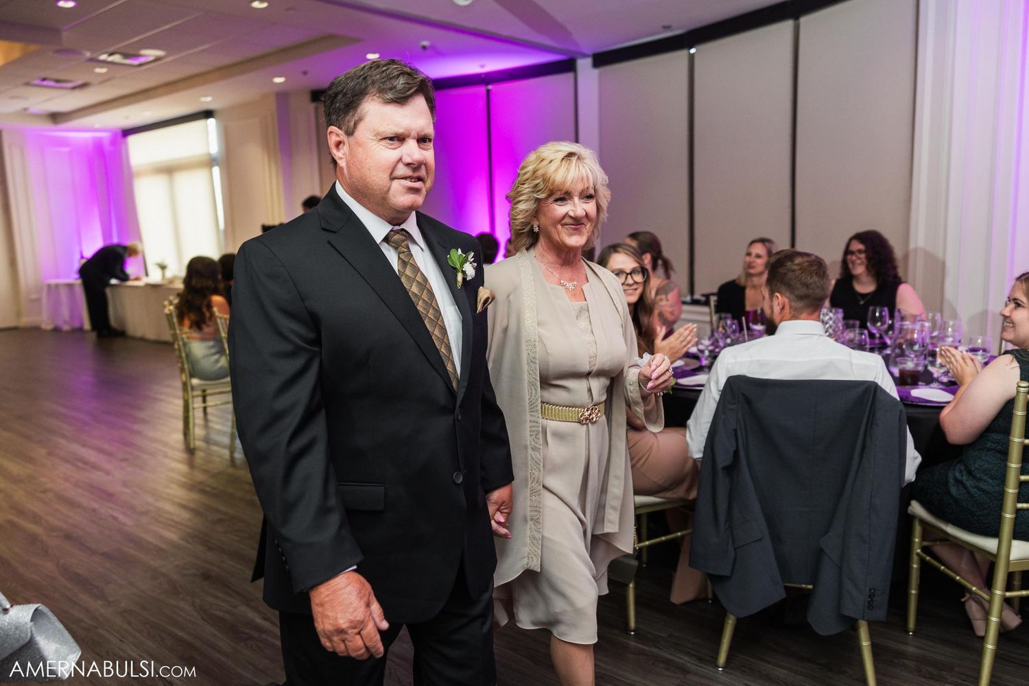 Wedding guests stand in an elegant reception hall with purple uplighting and formal table settings.