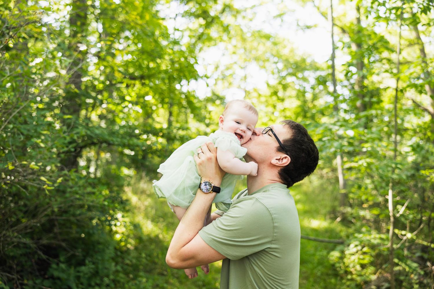 Joyful moment captured in sunlit forest as parent lifts baby wearing mint green dress overhead.
