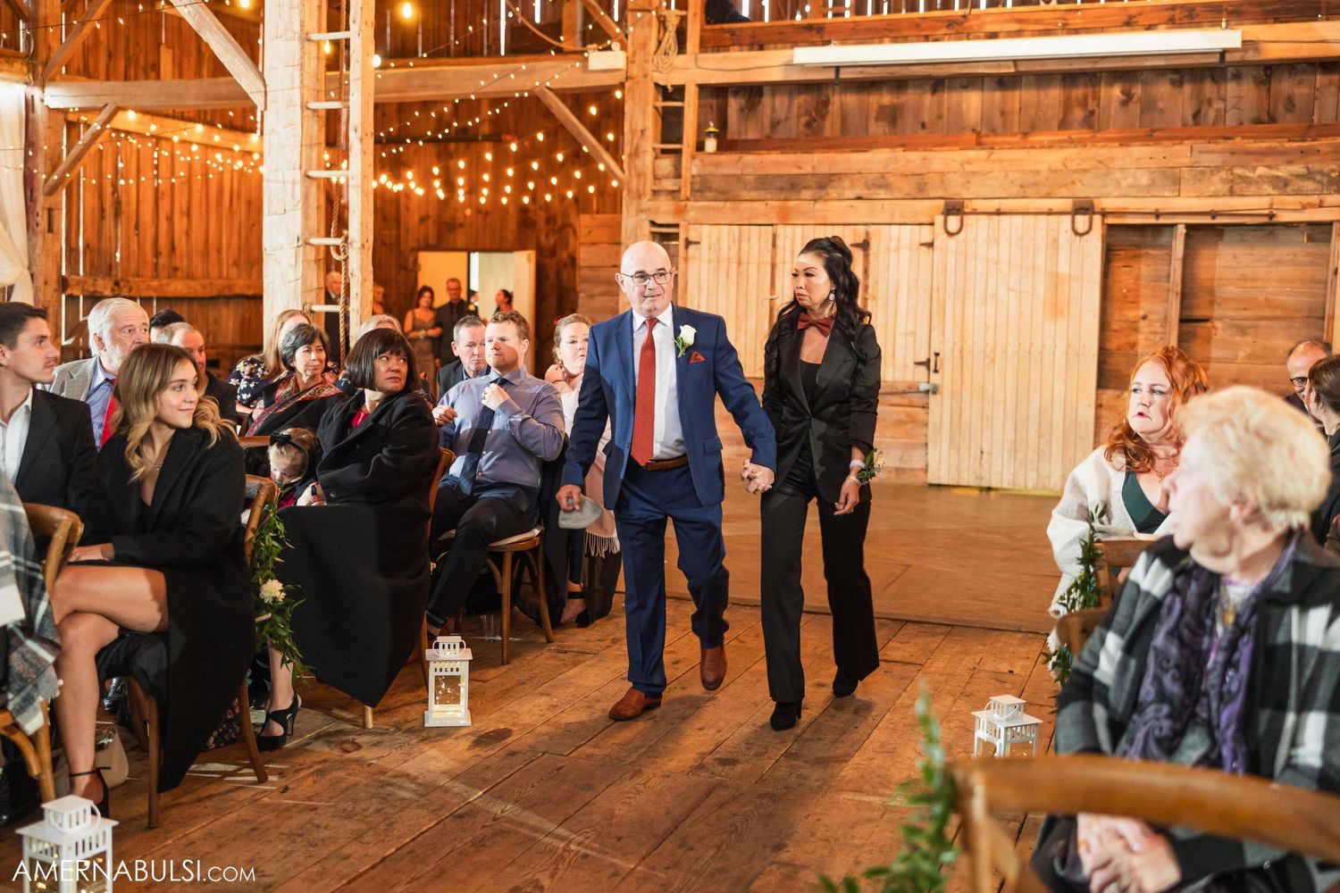 Wedding guests watch as attendees walk down the aisle in rustic barn venue with string lights.